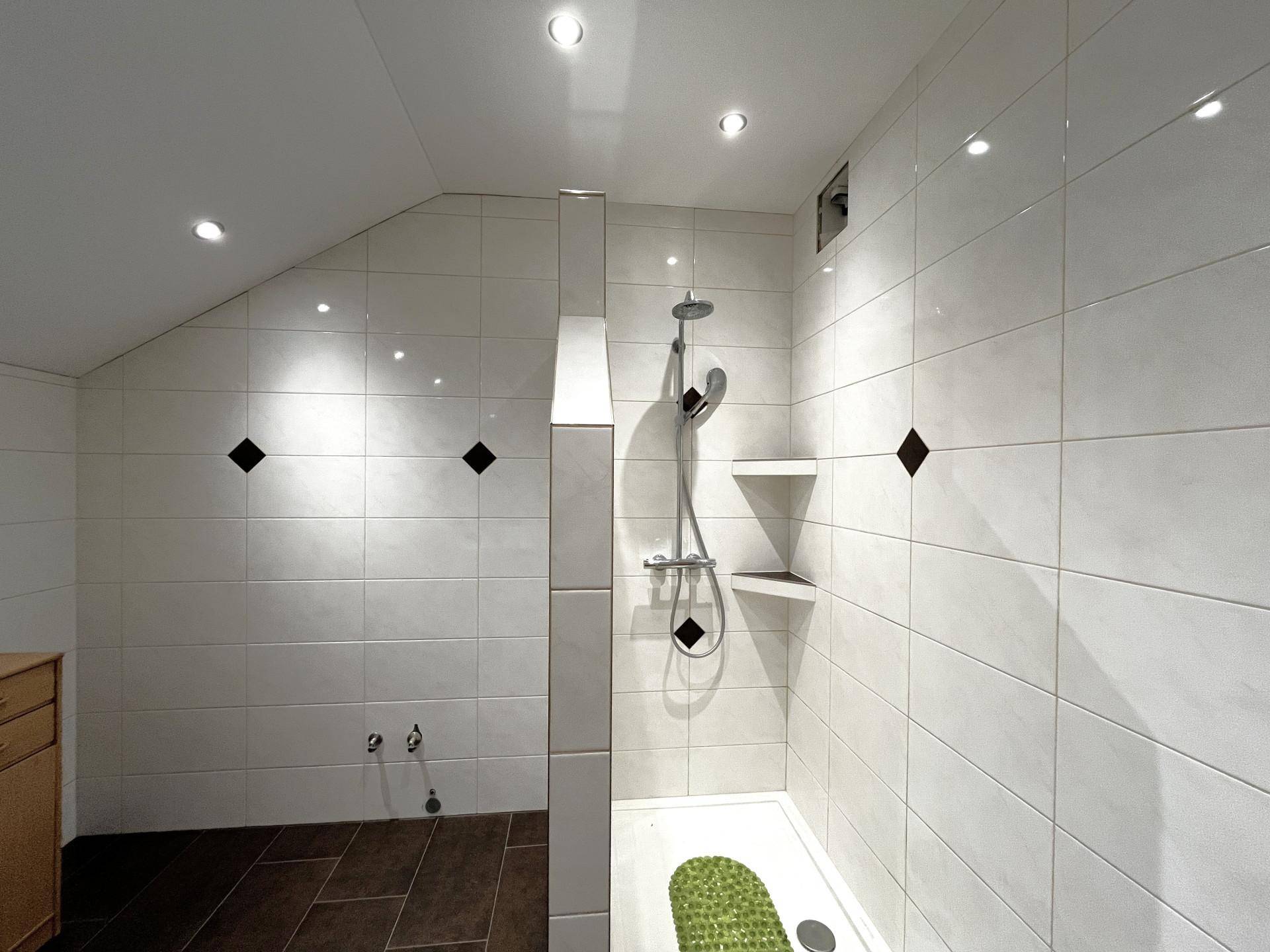 Duschbereich im Badezimmer mit weißen Fliesen, dunklem Boden und integrierter Ablage unter Dachschräge.