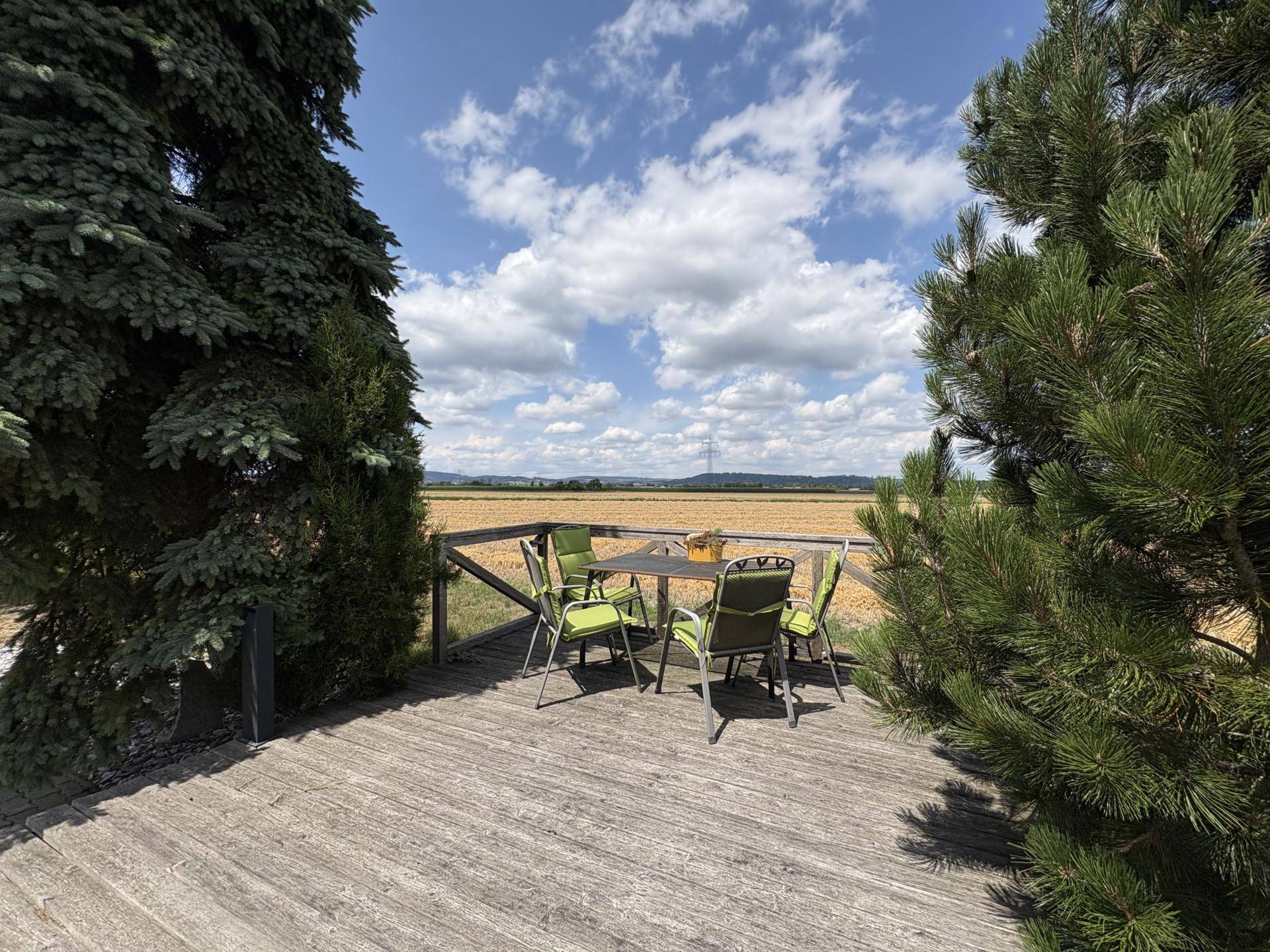 Holzterrasse mit Esstisch und Stühlen, umgeben von Bäumen mit weitem Blick auf die Landschaft.