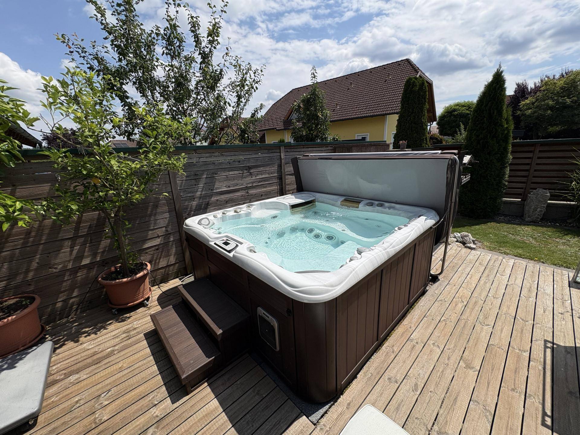 Außenwhirlpool auf einer Holzterrasse im Garten, umgeben von Pflanzen und Hecken.