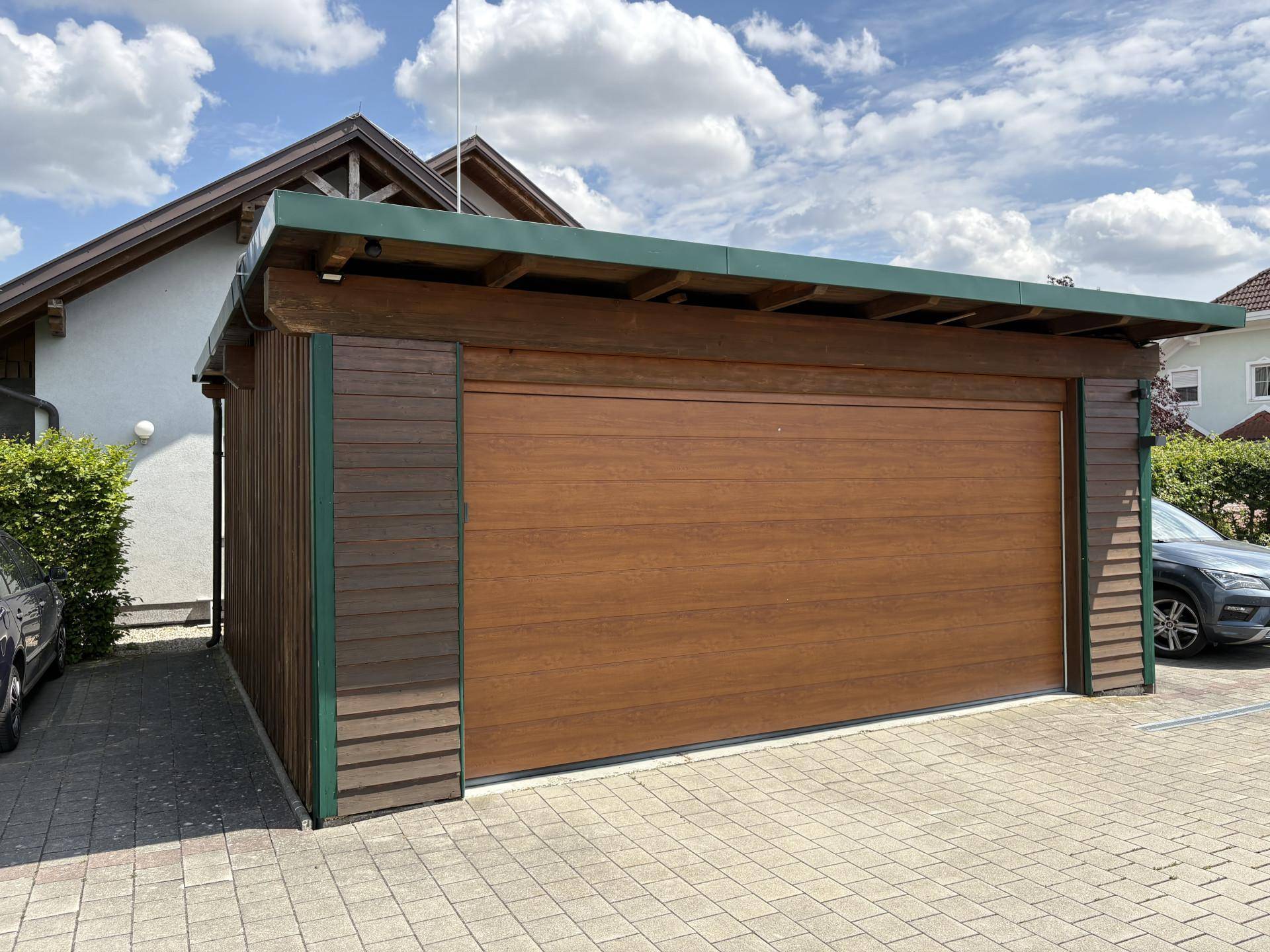 Einzelgarage aus Holz mit braunem Tor und grünem Dach, davor eine gepflasterte Einfahrt.