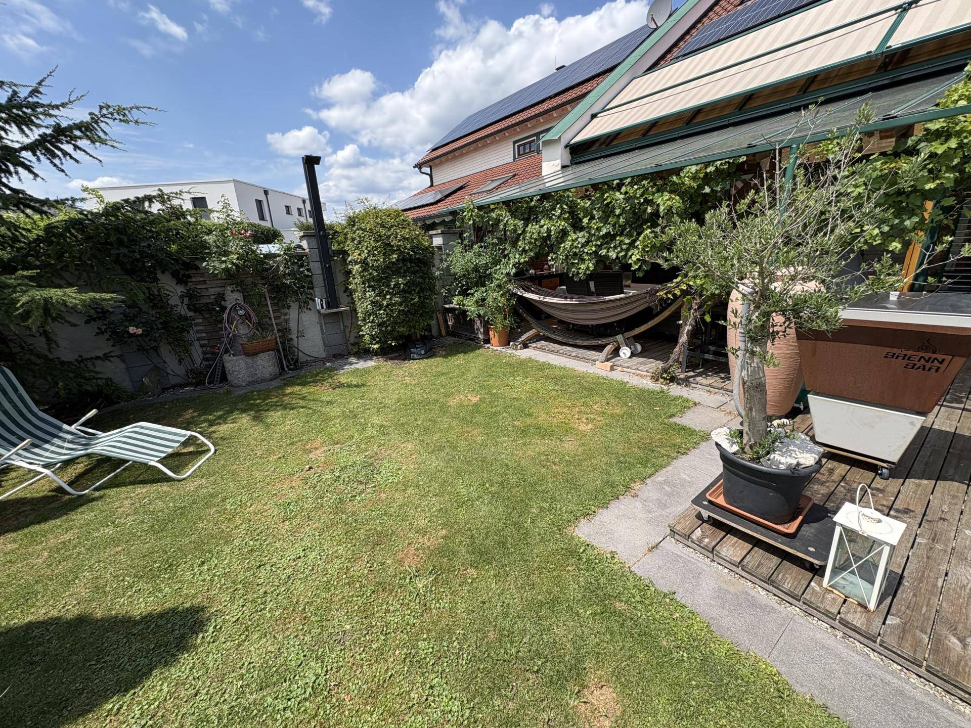 Grüner Gartenbereich mit Rasenfläche, Liegestuhl und Blick auf die überdachte Terrasse des Hauses.