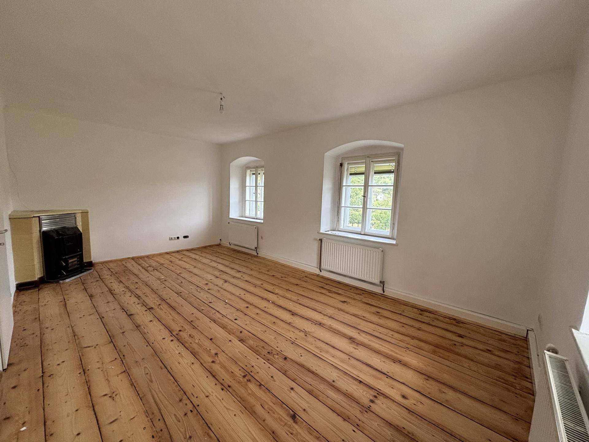 Helles, leeres Wohnzimmer mit breiten Holzplankenböden und einem Kaminofen in der Ecke.