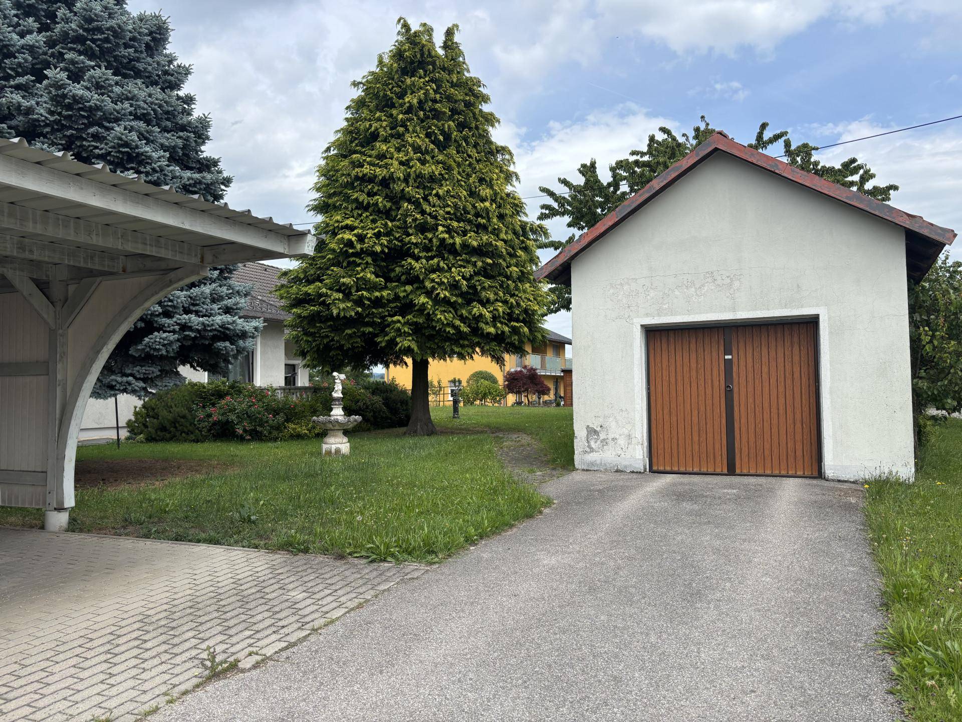Carport und separate Garage mit Holztor, umgeben von Rasen und Bäumen.
