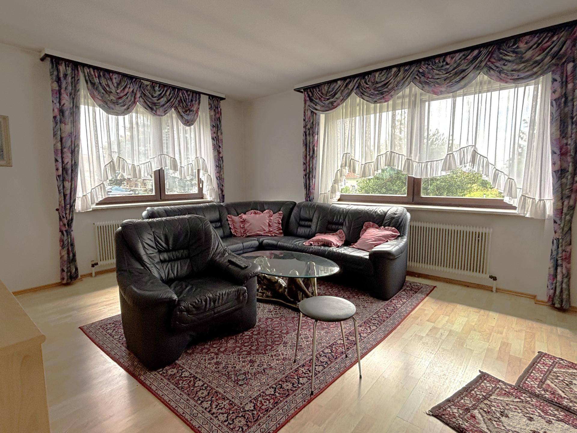 Gemütliches Wohnzimmer mit Ledersofa, Teppich und großen Fenstern mit Vorhängen.