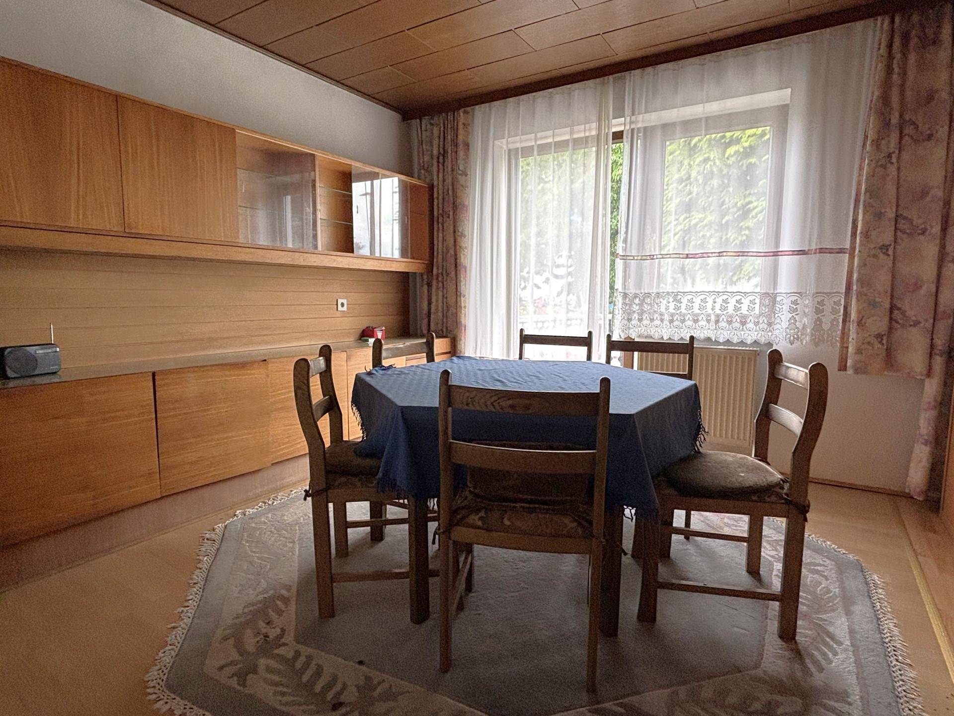 Esszimmer mit Holztisch, Stühlen und Holzschränken, durch eine große Fenstertür hell beleuchtet.