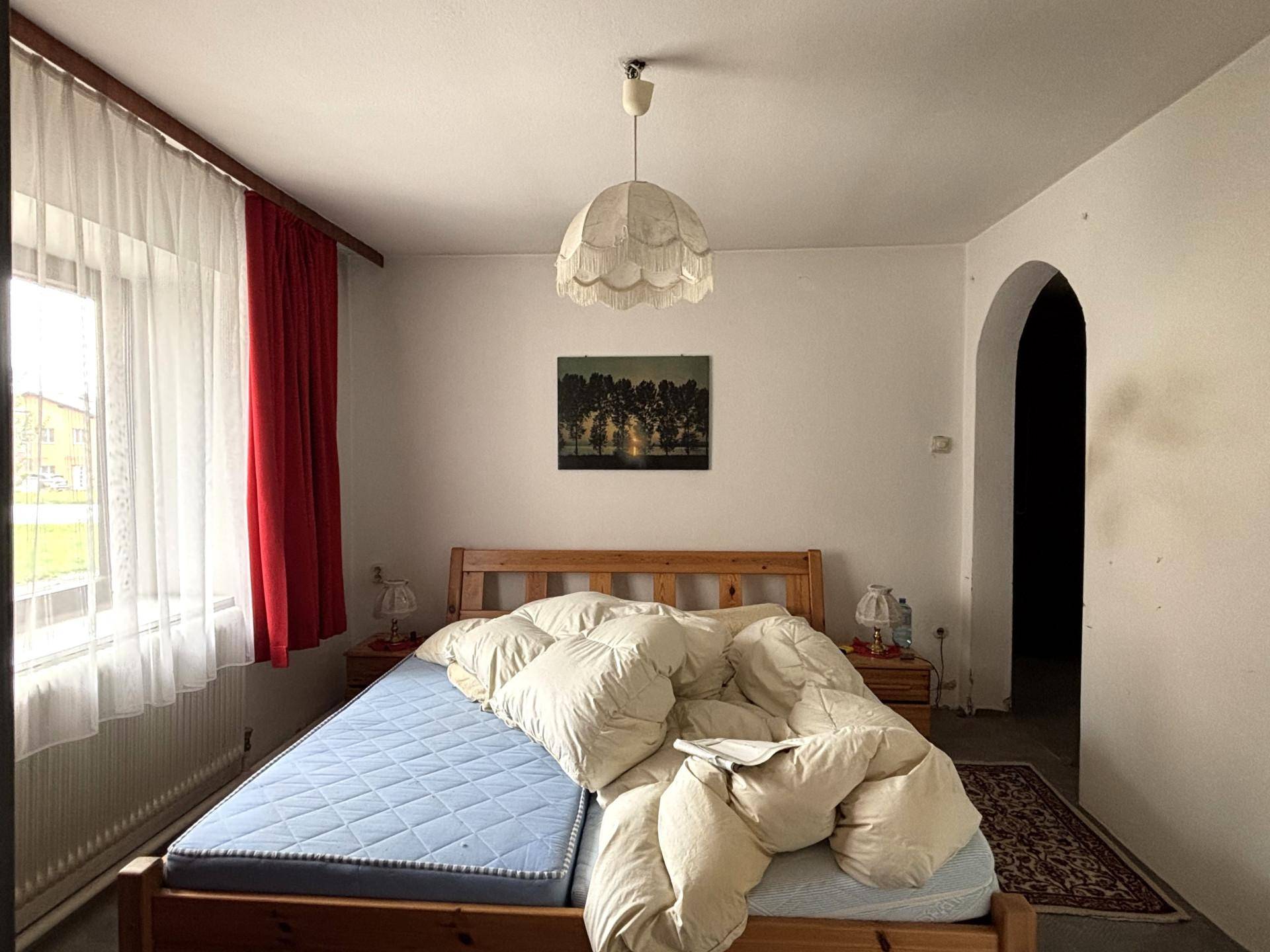Schlafzimmer mit Holzbett, einfacher Einrichtung und einem Fenster mit rotem Vorhang.