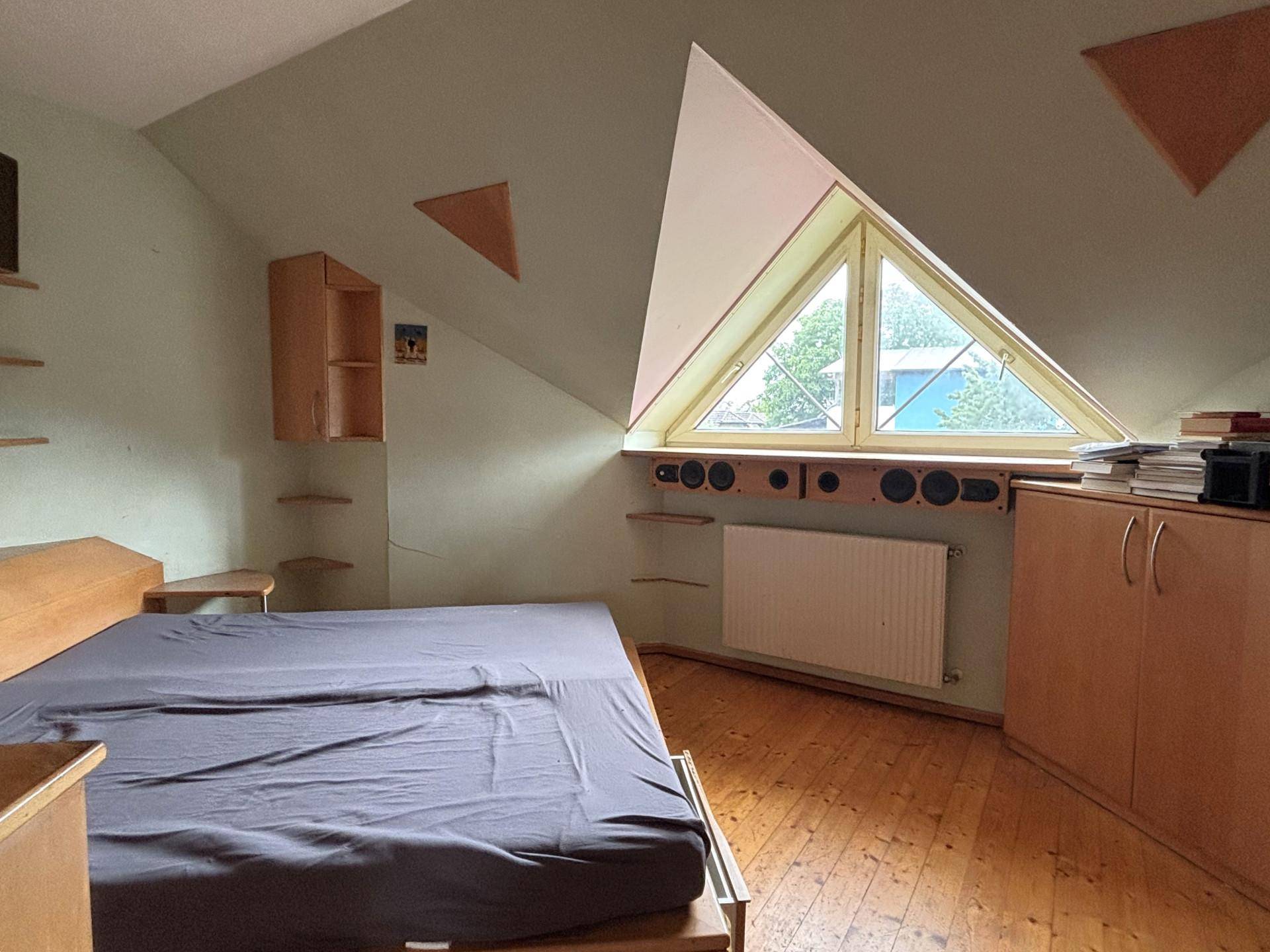 Schlafzimmer im Dachgeschoss mit Holzboden, schrägen Wänden und einem dreieckigen Fenster.