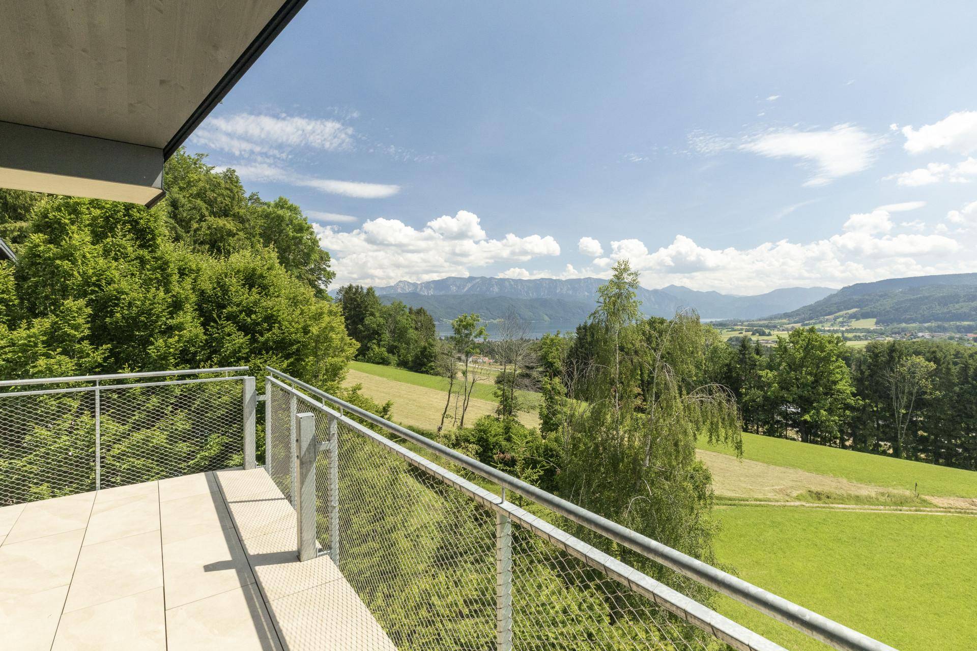 Geräumiger Balkon mit Fliesenboden und weitem Blick über grüne Landschaft, See und Berge.