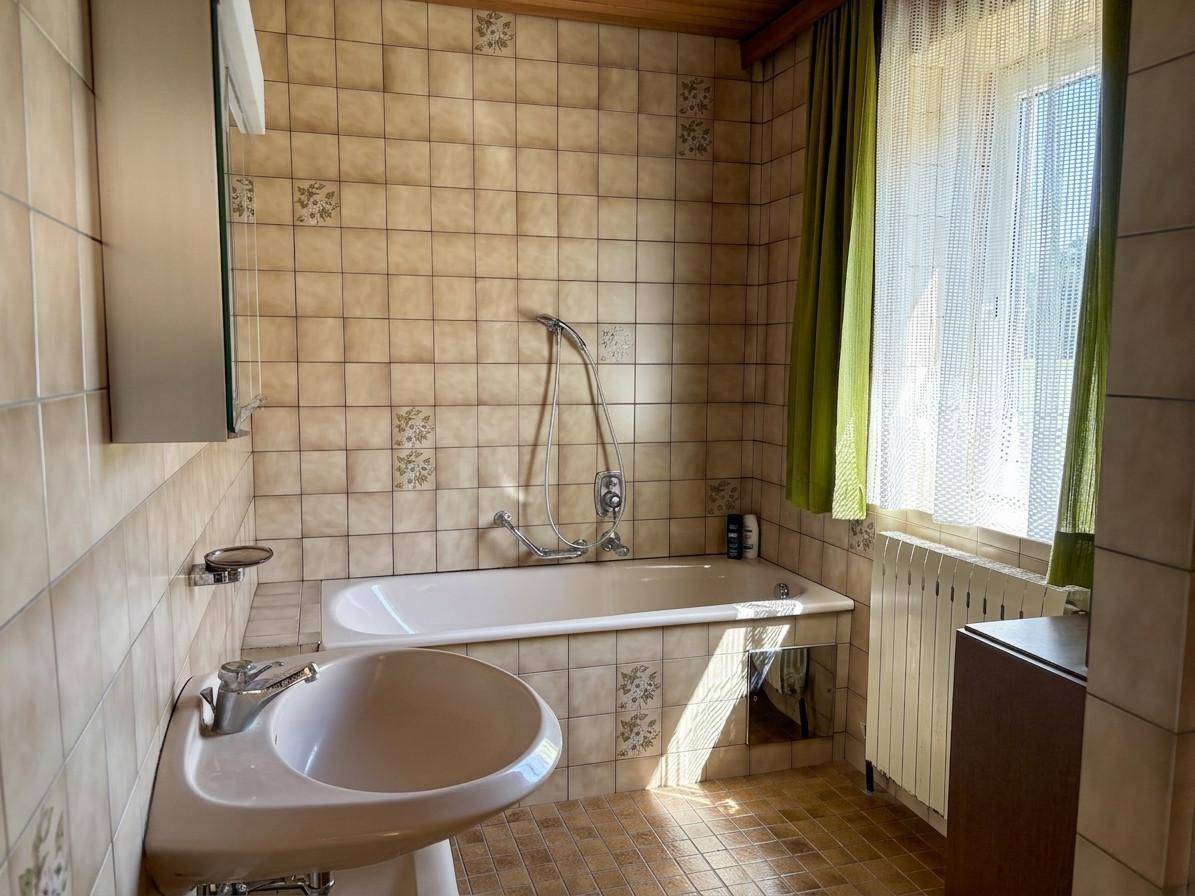 Gefliestes Badezimmer mit Badewanne, Waschbecken und Fenster, im Stil der 80er Jahre.
