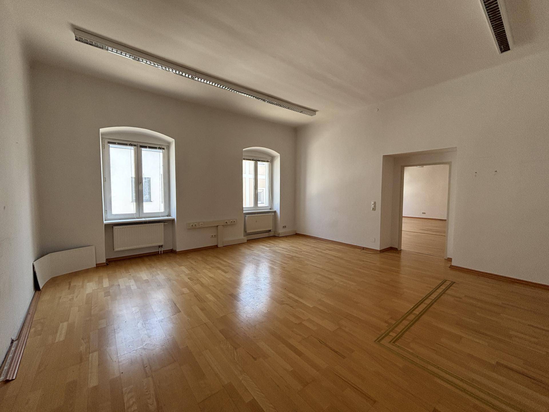 Heller Büroraum mit Parkettboden, hohen Decken und zwei Fenstern, die den Raum gut belichten.