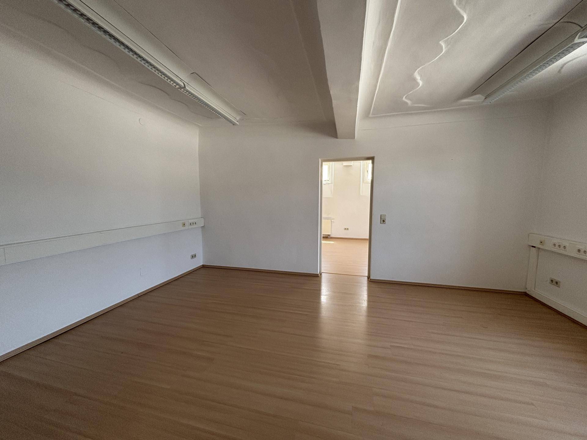 Leerer Büroraum mit hohen Decken, Holzboden und einem Durchgang zu einem weiteren Zimmer, gut beleuchtet.