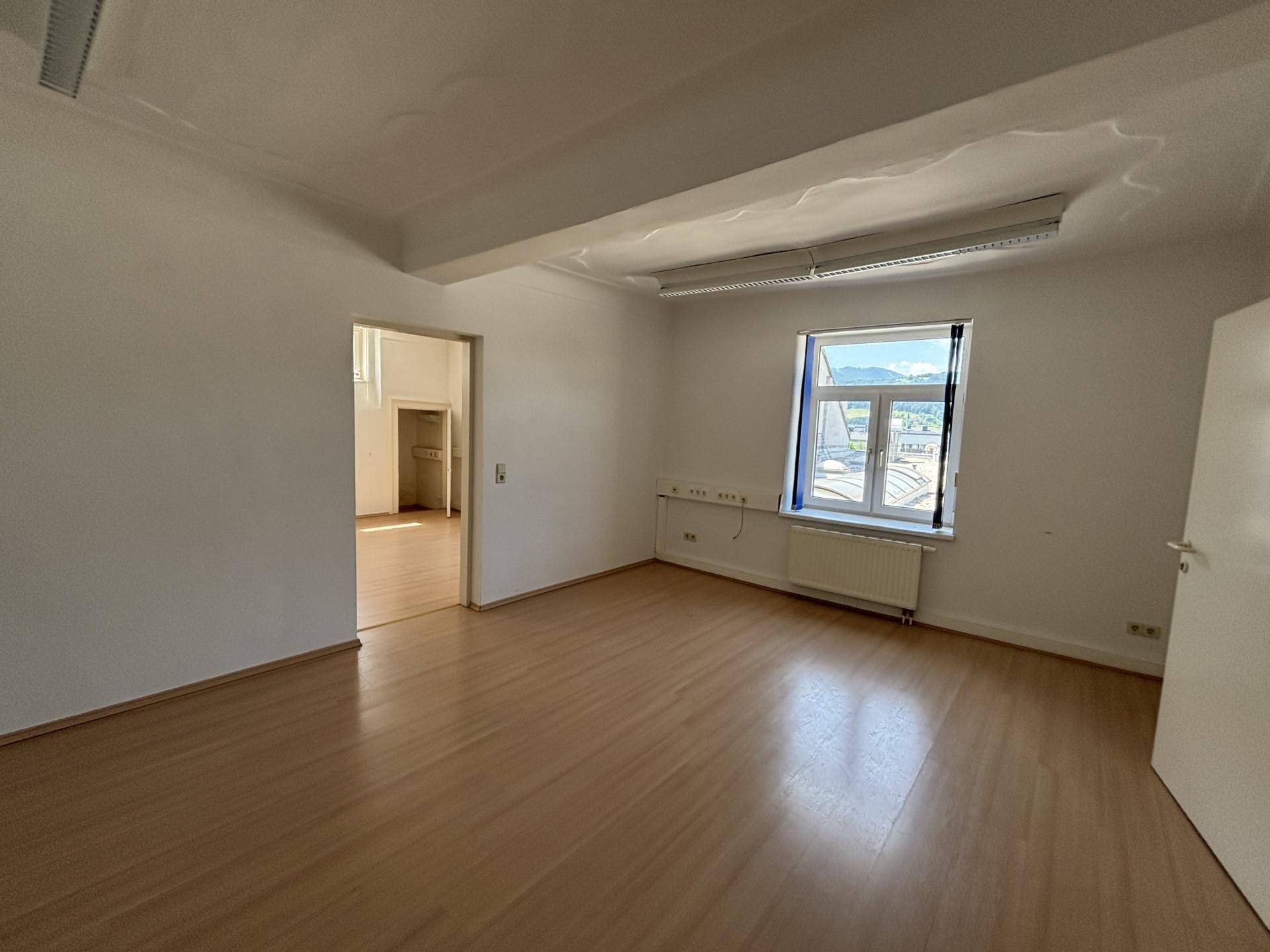 Heller Büroraum mit Holzboden, hohen Decken und einem Fenster, das einen weiten Blick ermöglicht.