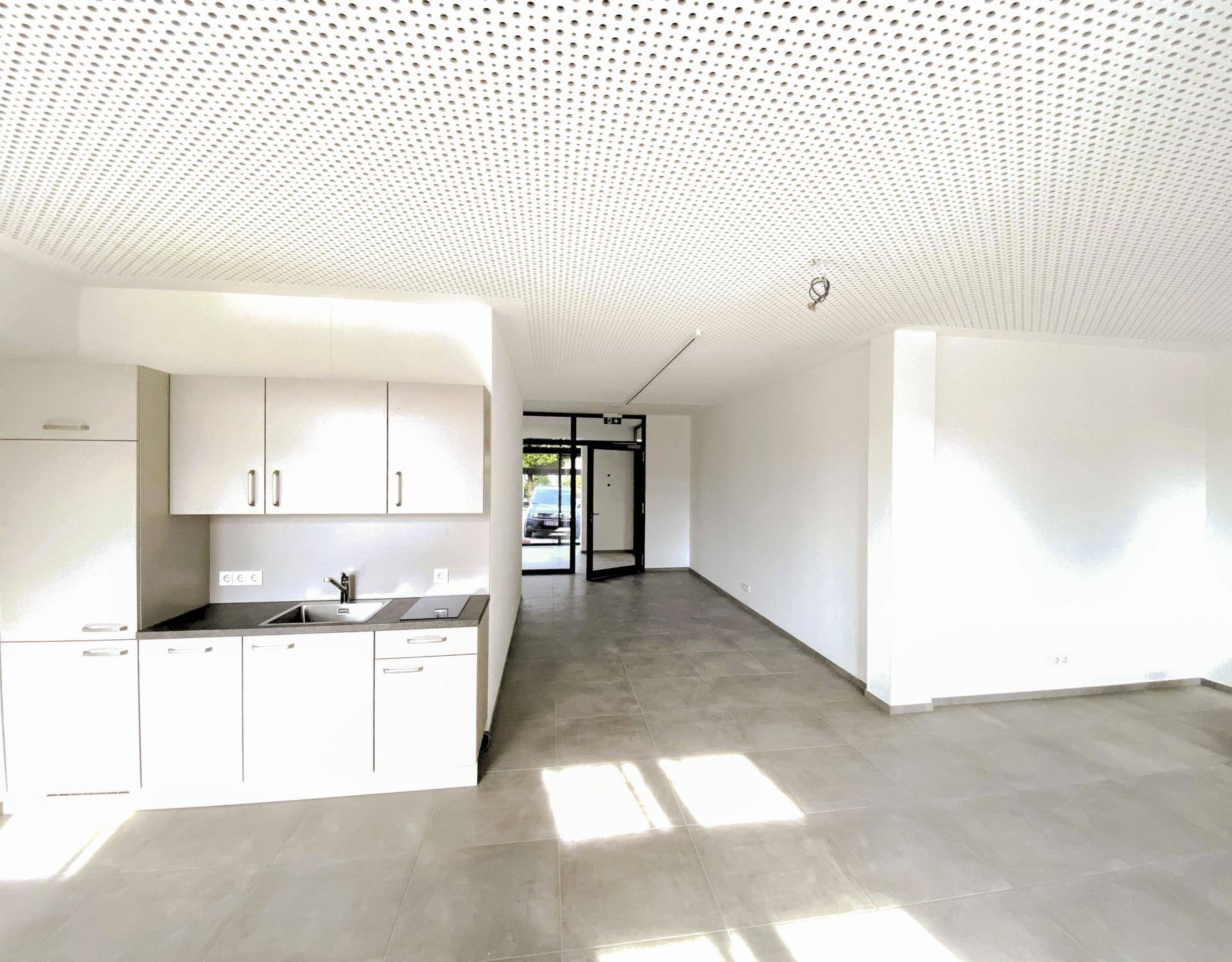 Moderne Küchenzeile in einem hellen Gewerberaum mit weißen Schränken, Induktionskochfeld und grauen Bodenfliesen.