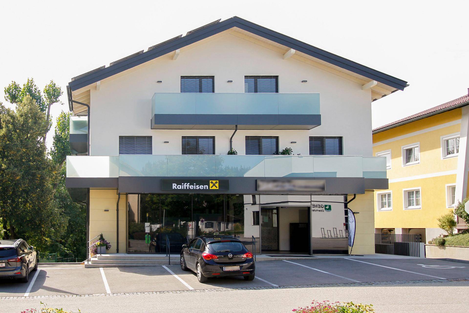 Modernes Geschäftsgebäude mit Glasbalkonen, Raiffeisen-Filiale im Erdgeschoss und Parkplätzen davor.