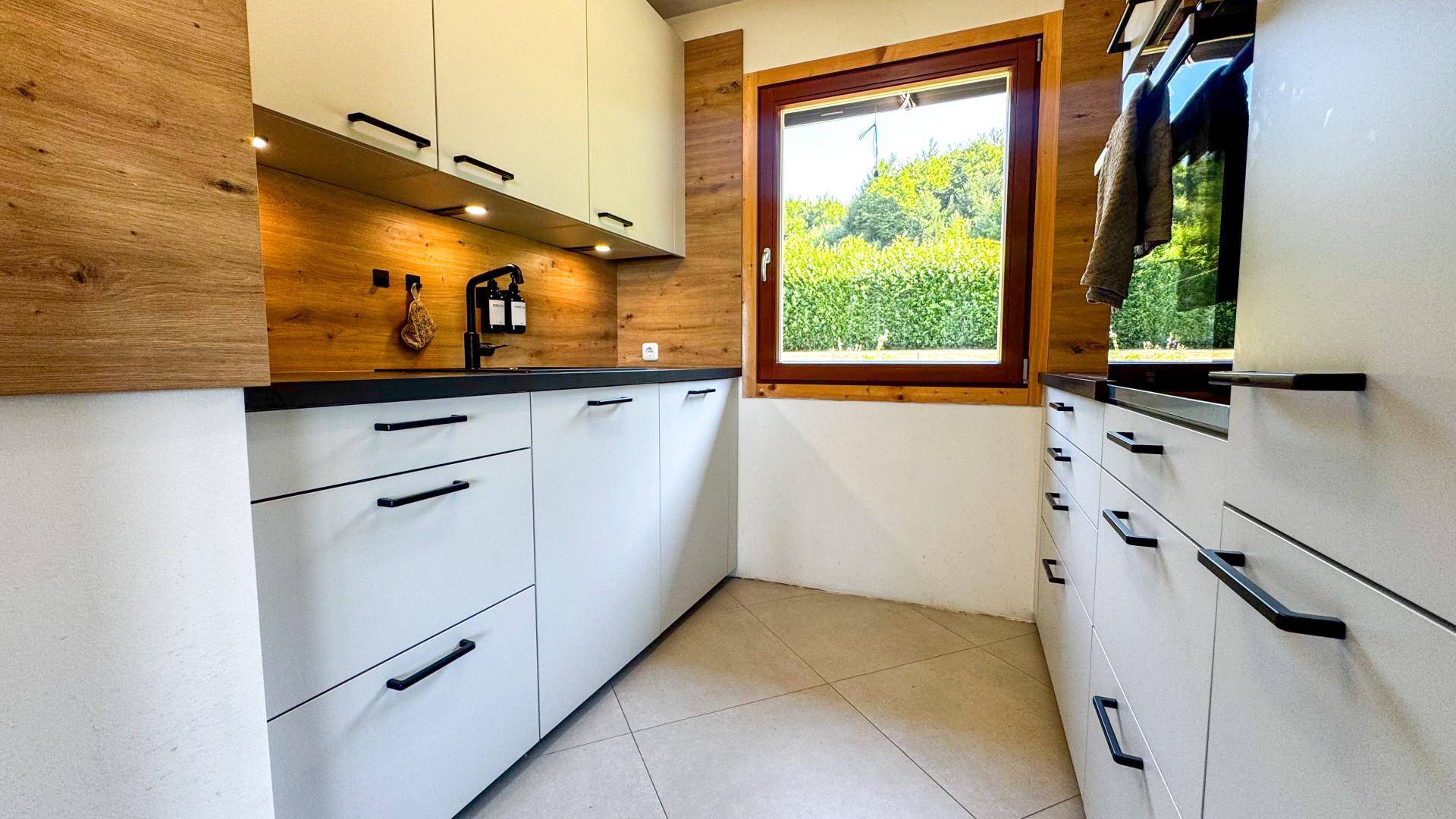 Moderne Küche mit weißen Schränken, Holzakzenten und Blick ins Grüne durch das Fenster.