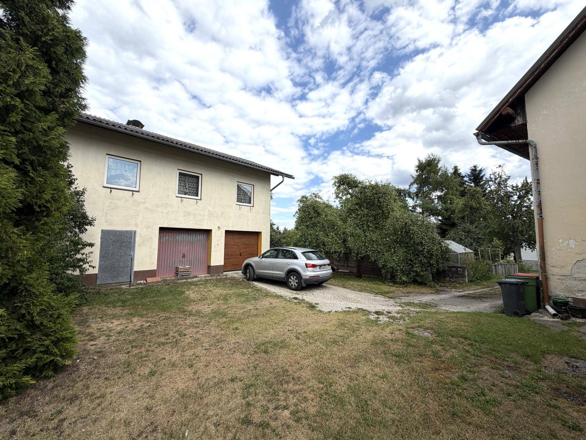 Rückseite des Hauses mit zwei Garagen und einem Kiesweg, umgeben von Bäumen.