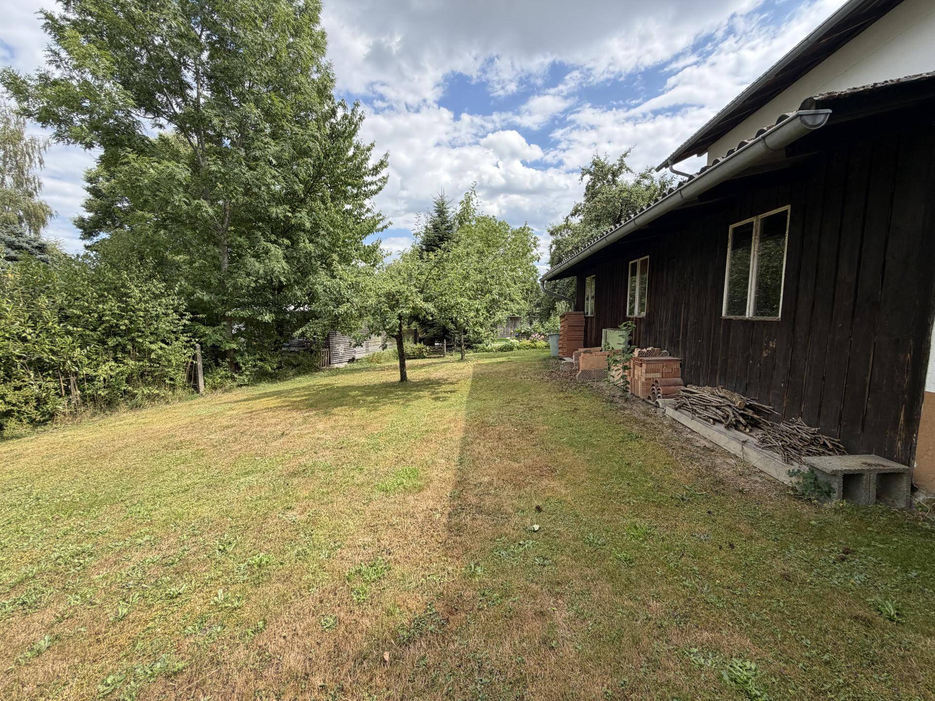 Großer Gartenbereich mit Rasenfläche und altem Baumbestand neben der Holzfassade des Hauses unter bewölktem Himmel.