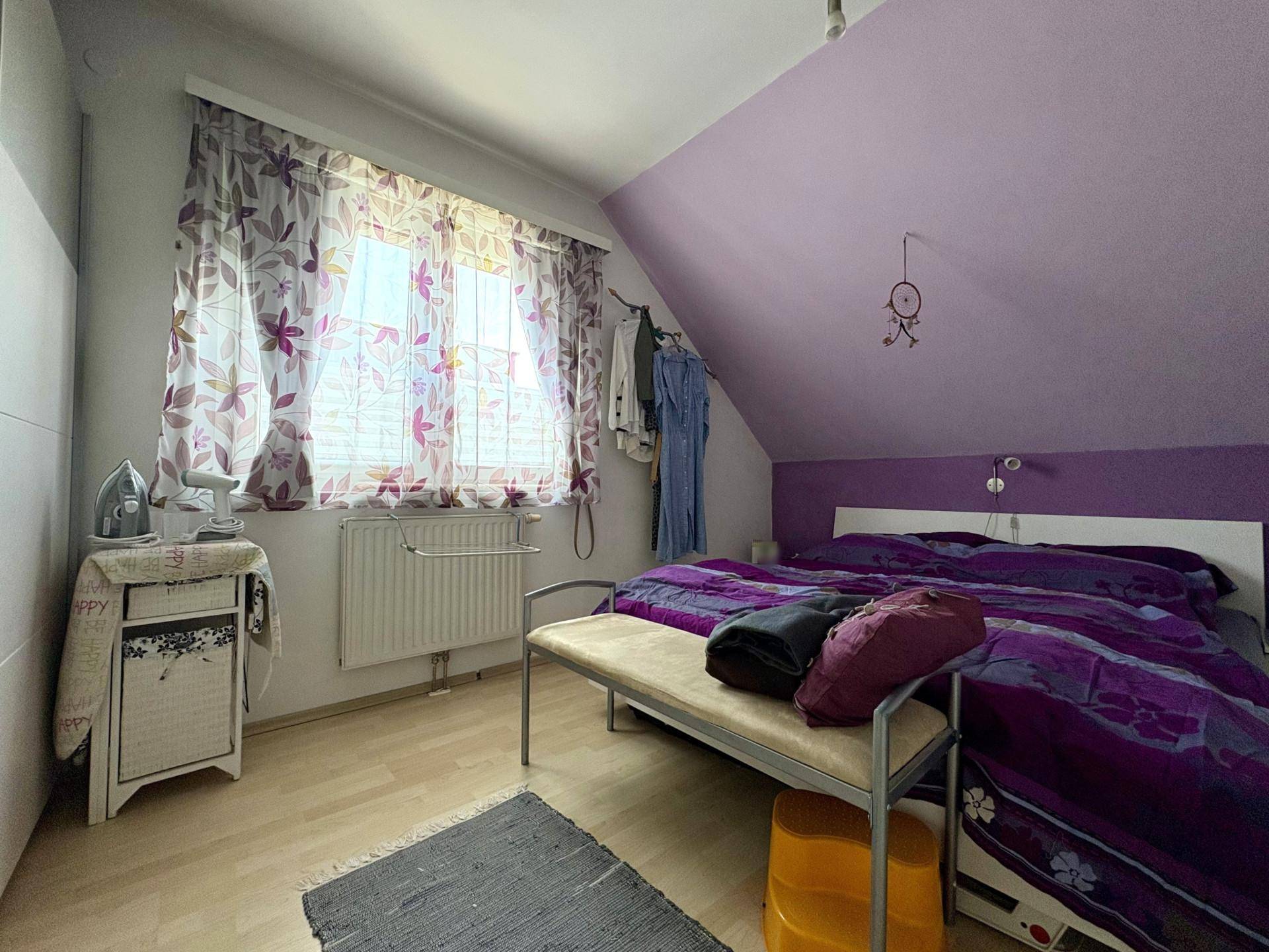 Schlafzimmer im Dachgeschoss mit Bett, Fenster und lila gestrichener Wand.