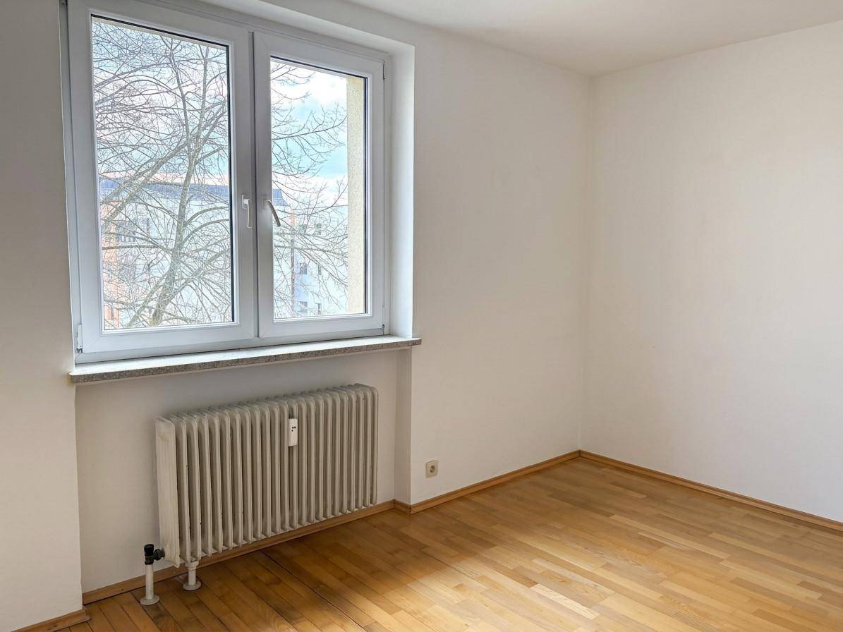 Leeres Zimmer mit Holzboden, weißen Wänden und einem großen Fenster mit Blick ins Grüne.