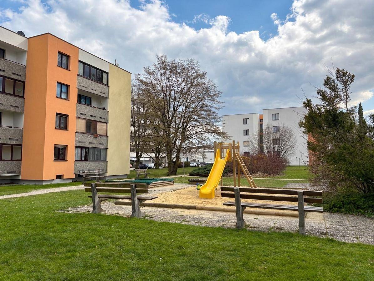 Grüner Gemeinschaftsgarten mit Spielplatz, Rutsche und Sitzbänken vor einem Mehrfamilienhaus.