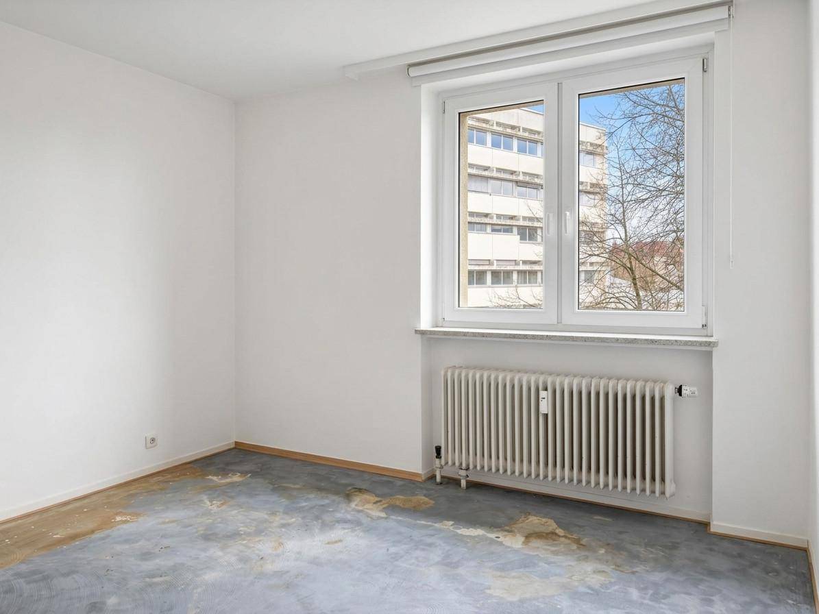 Unmöbliertes Zimmer mit beschädigtem Boden, weißen Wänden und Fensterblick auf andere Gebäude.