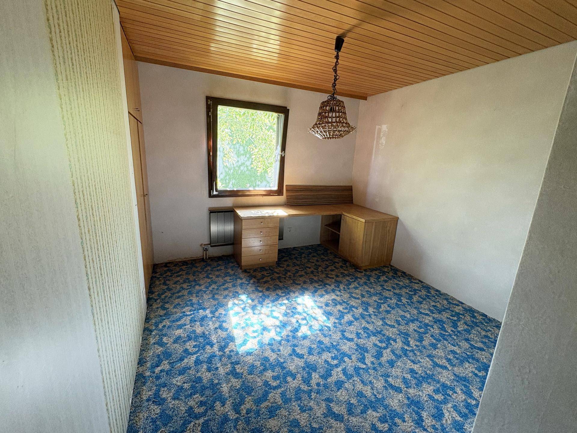 Helles Arbeitszimmer mit blau gemustertem Teppichboden, Holzschreibtisch und Holzpaneeldecke.