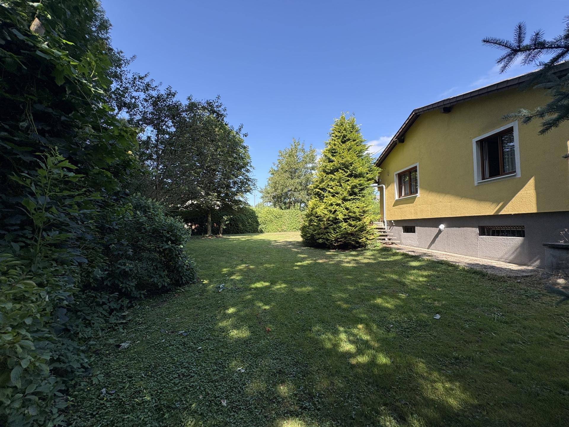 Seitenansicht des Hauses mit weitläufigem Rasen und hohen Bäumen unter blauem Himmel.