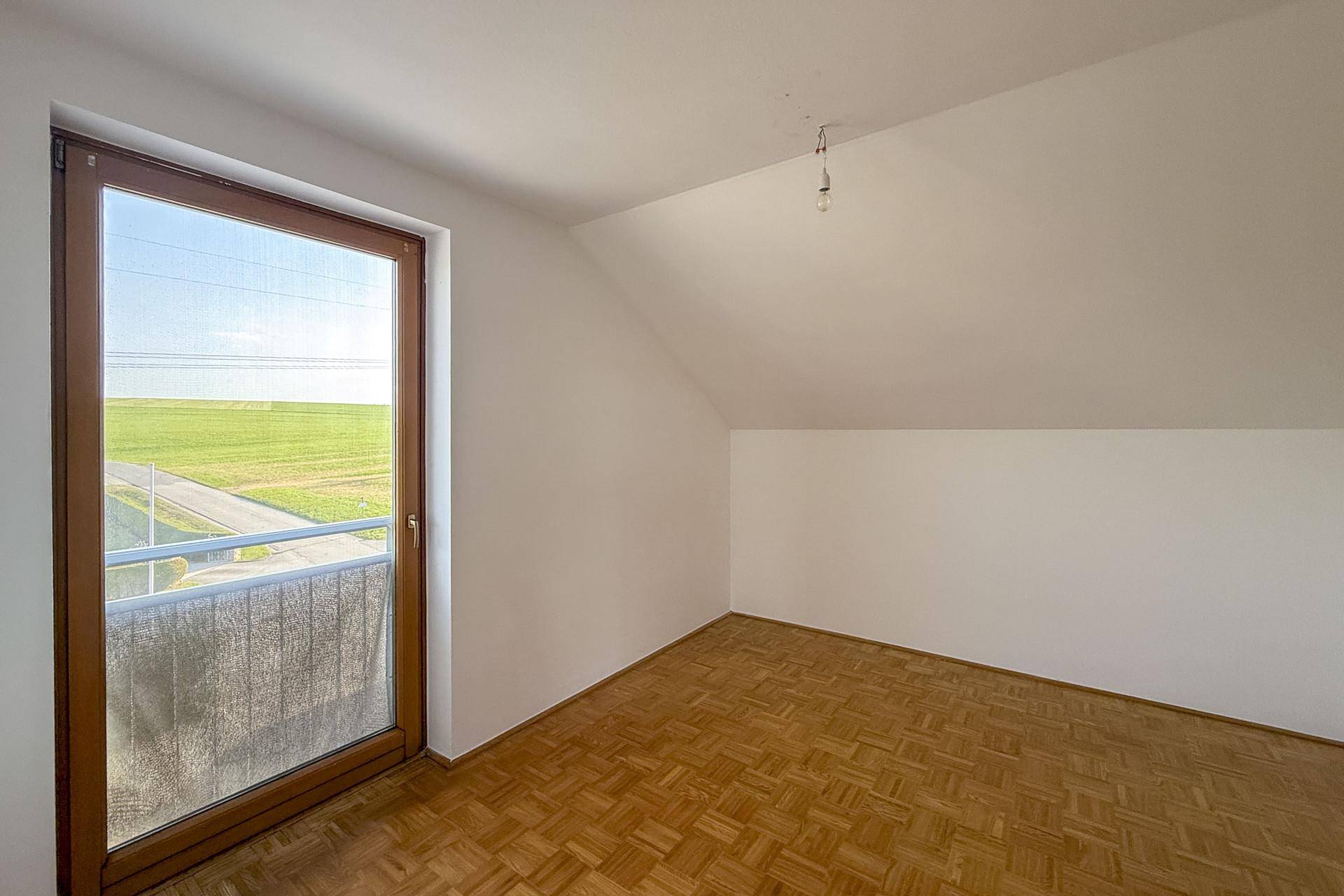 Gemütliches Zimmer mit Parkettboden, Dachschräge und Zugang zu einem kleinen Balkon.