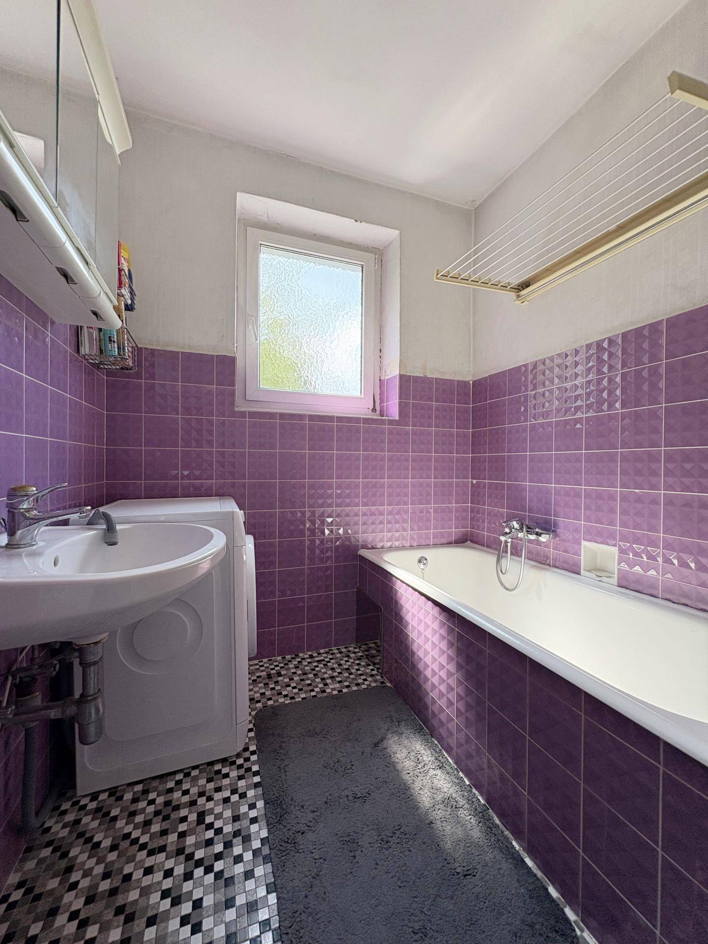 Badezimmer mit lila Fliesen, Badewanne, Waschmaschine und Fenster für natürliche Belüftung.
