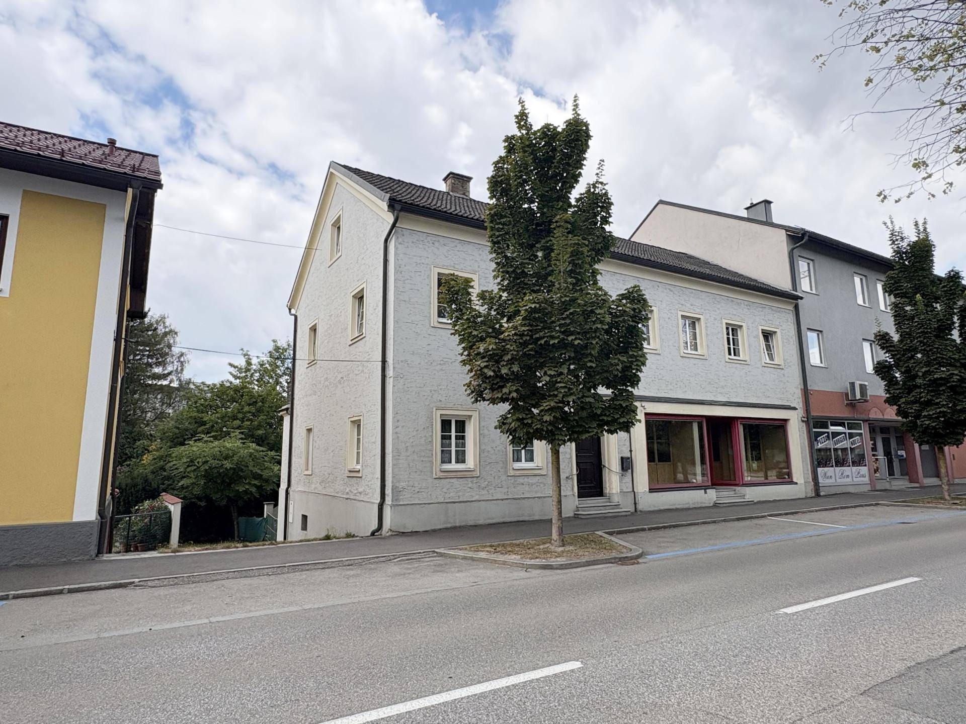 Seitenansicht des Gebäudes mit Giebeldach und weiteren Fenstern, umgeben von Bäumen und einer Straße.