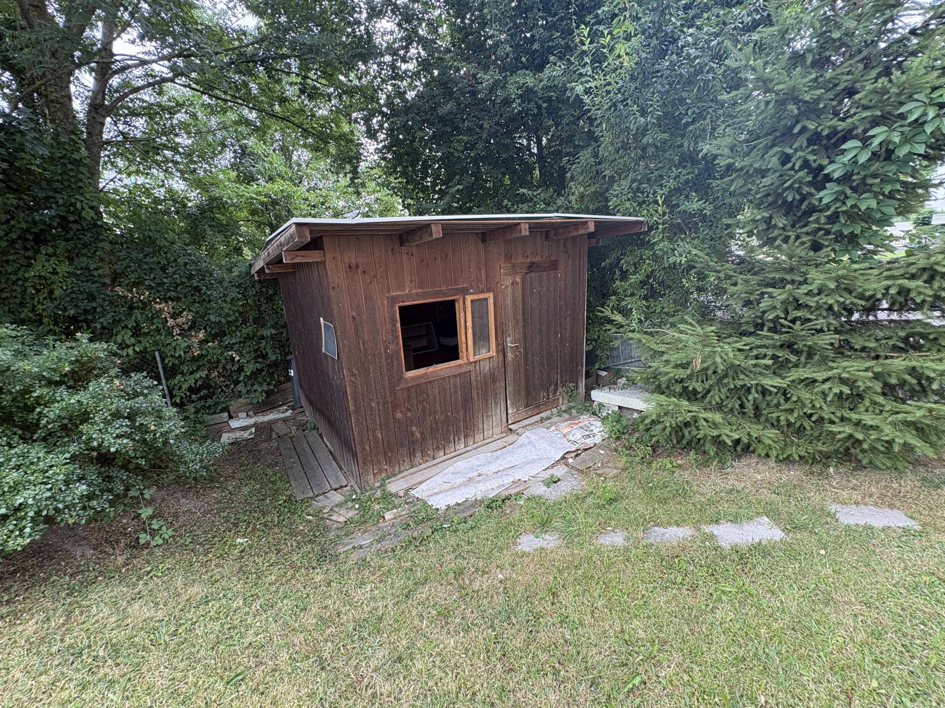 Kleiner hölzerner Geräteschuppen mit Fenster, eingebettet in eine grüne Gartenlandschaft.