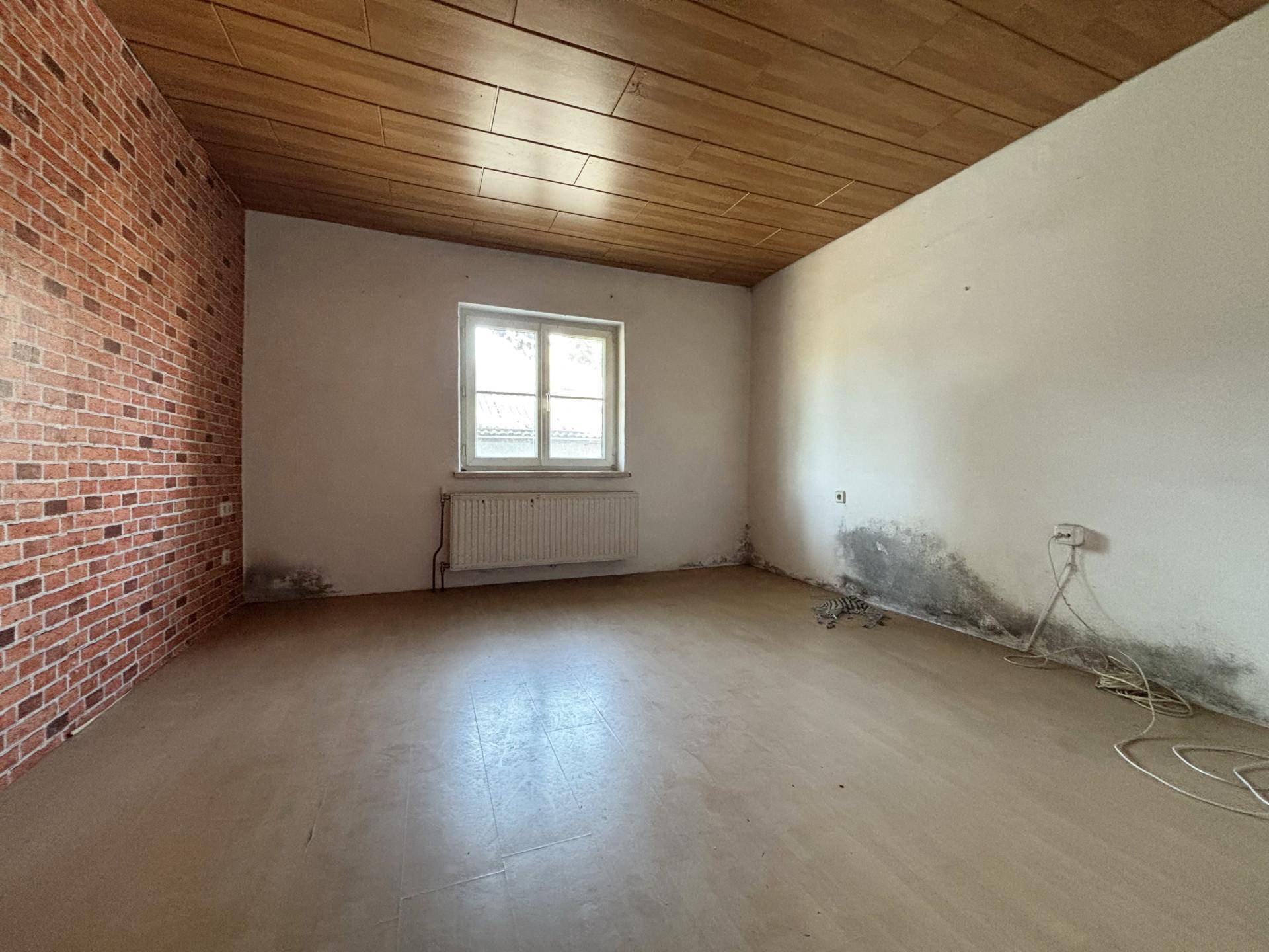 Raum mit Ziegelwandmuster, Holzdecke, Fenster und Heizkörper, zeigt Feuchtigkeitsschäden.
