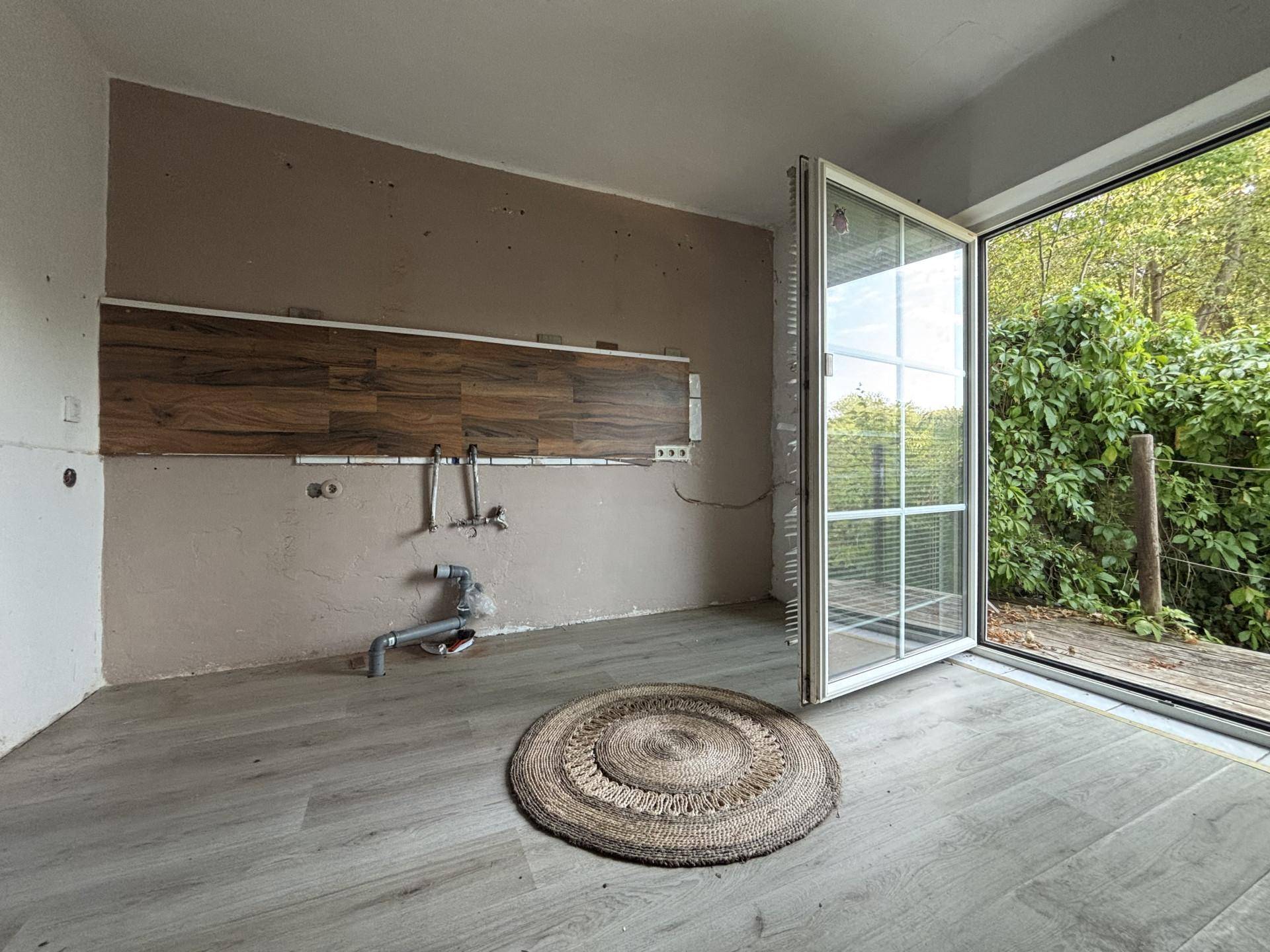 Leerer Küchenraum mit Anschlüssen, Holzwandverkleidung und großem Fenster zum Außenbereich.