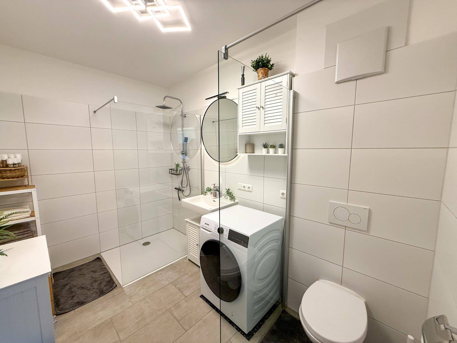 Modernes Badezimmer mit ebenerdiger Dusche, Waschmaschine und weißen Fliesen.