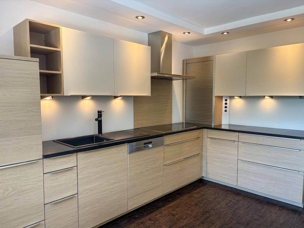 Moderne Einbauküche mit hellen Holzfronten, dunklen Arbeitsplatten, integrierten Geräten und indirekter Beleuchtung.