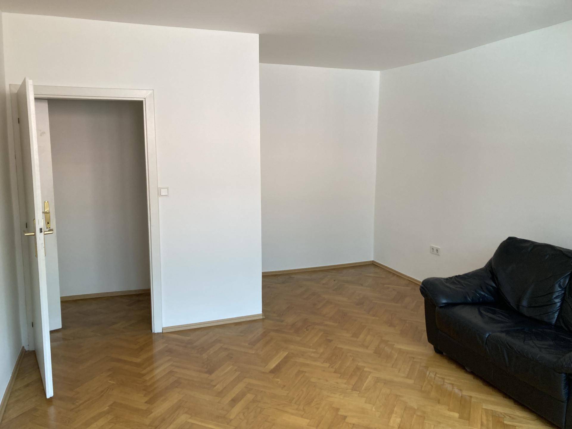 Geräumiges Wohnzimmer mit Fischgrätparkett und einer schwarzen Ledercouch.