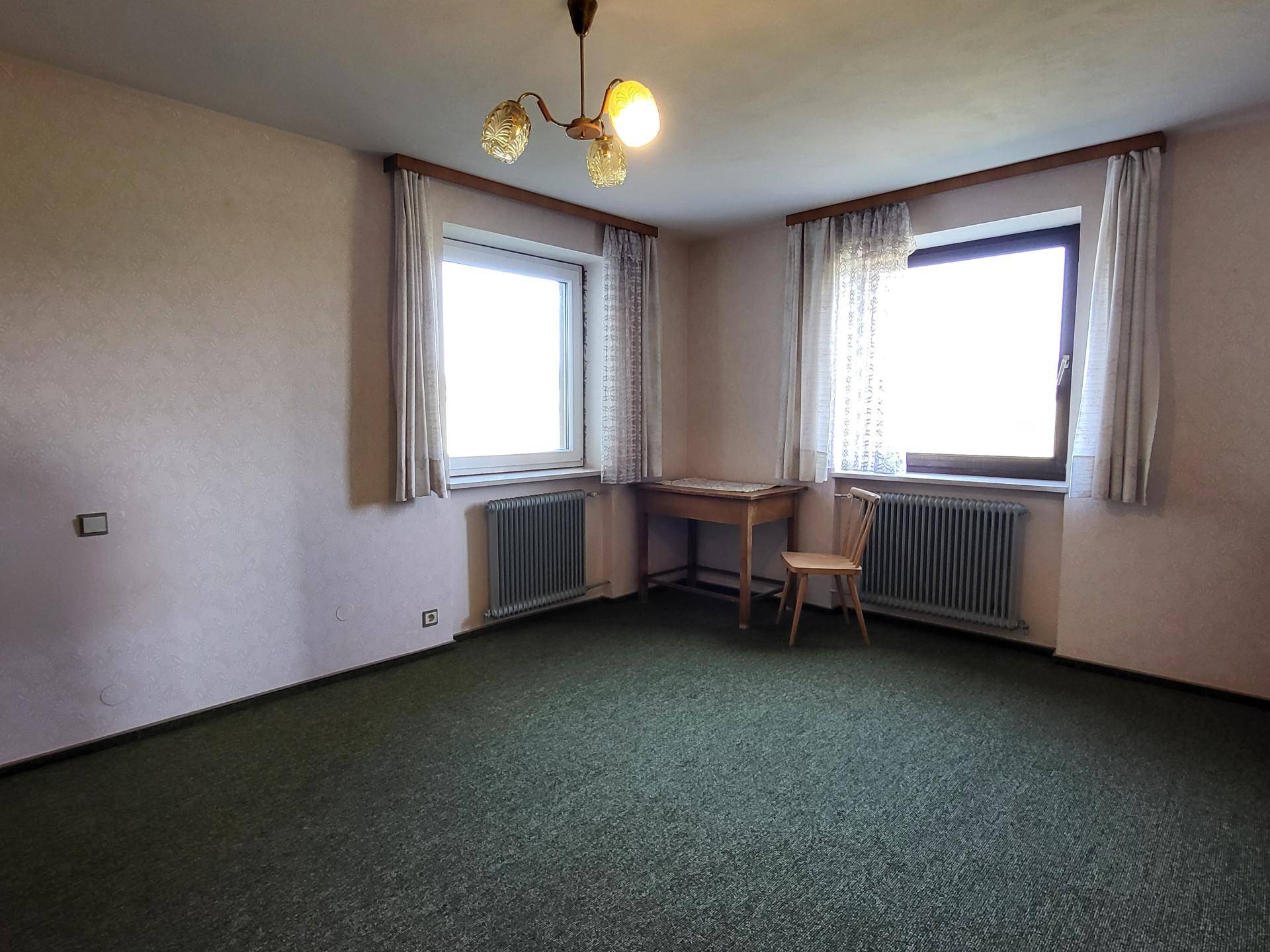 Schlafzimmer mit Teppichboden, zwei Fenstern und einfacher Einrichtung.