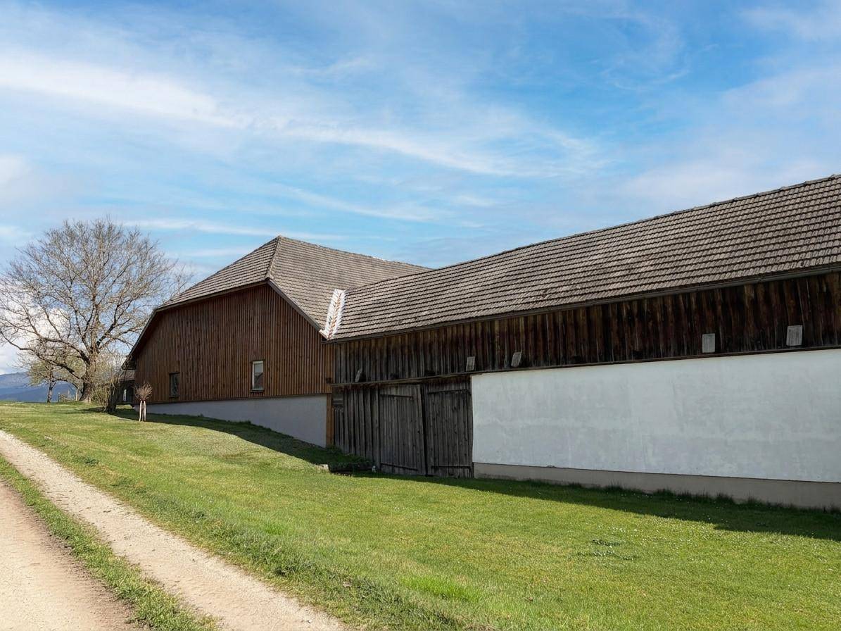 Ländliches Gebäude mit Holzwänden und Ziegeldach unter blauem Himmel, umgeben von Grünflächen.