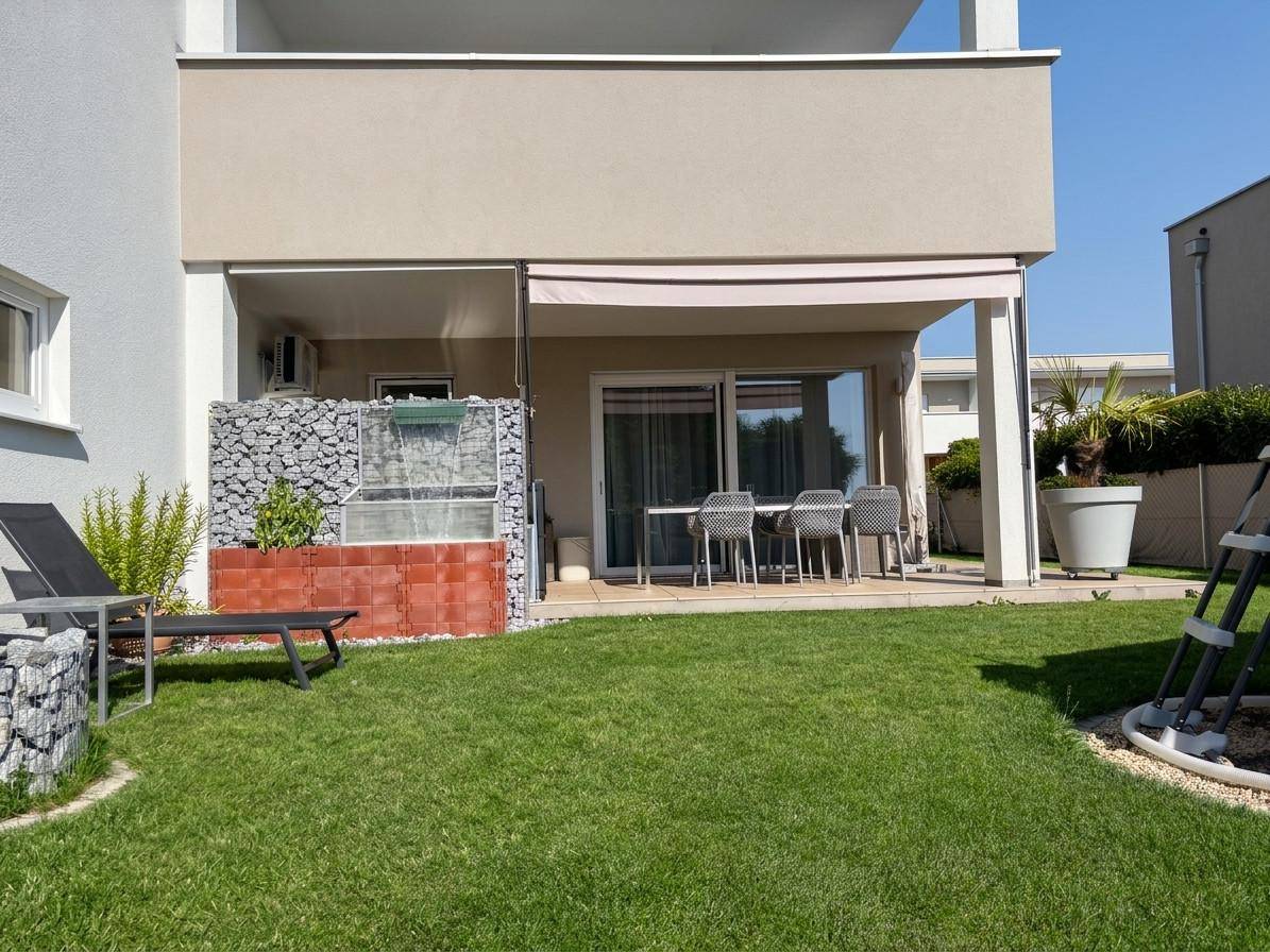 Überdachte Terrasse mit Sitzgelegenheiten und einem kleinen Wasserfall im sonnigen Gartenbereich.