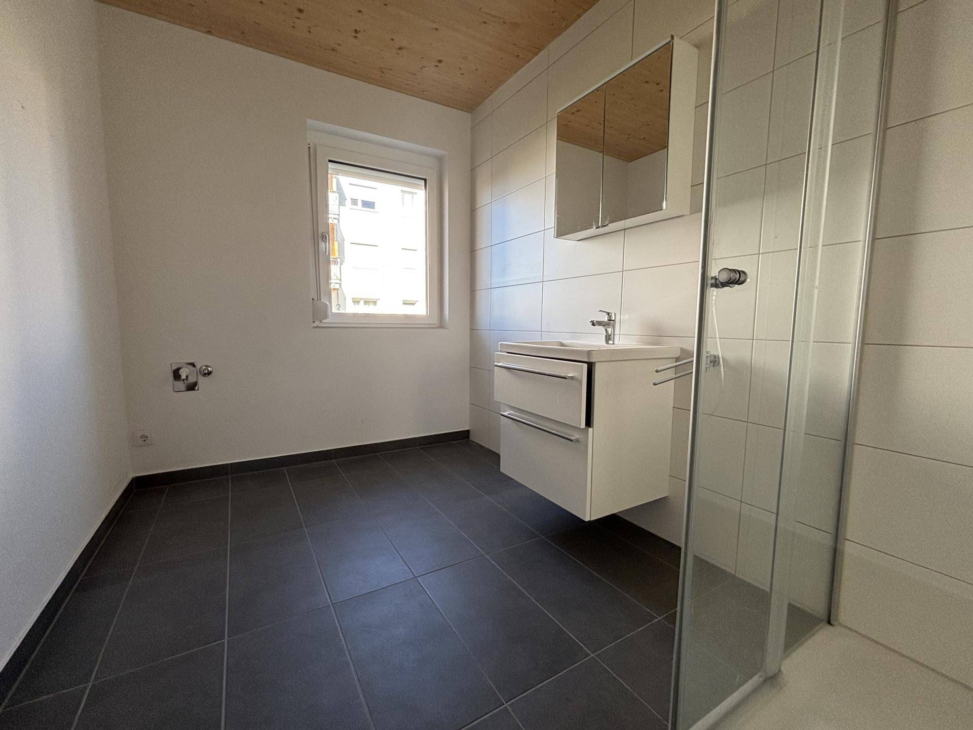 Helles Badezimmer mit Fenster, Duschkabine und Waschtisch, Platz für Waschmaschine.