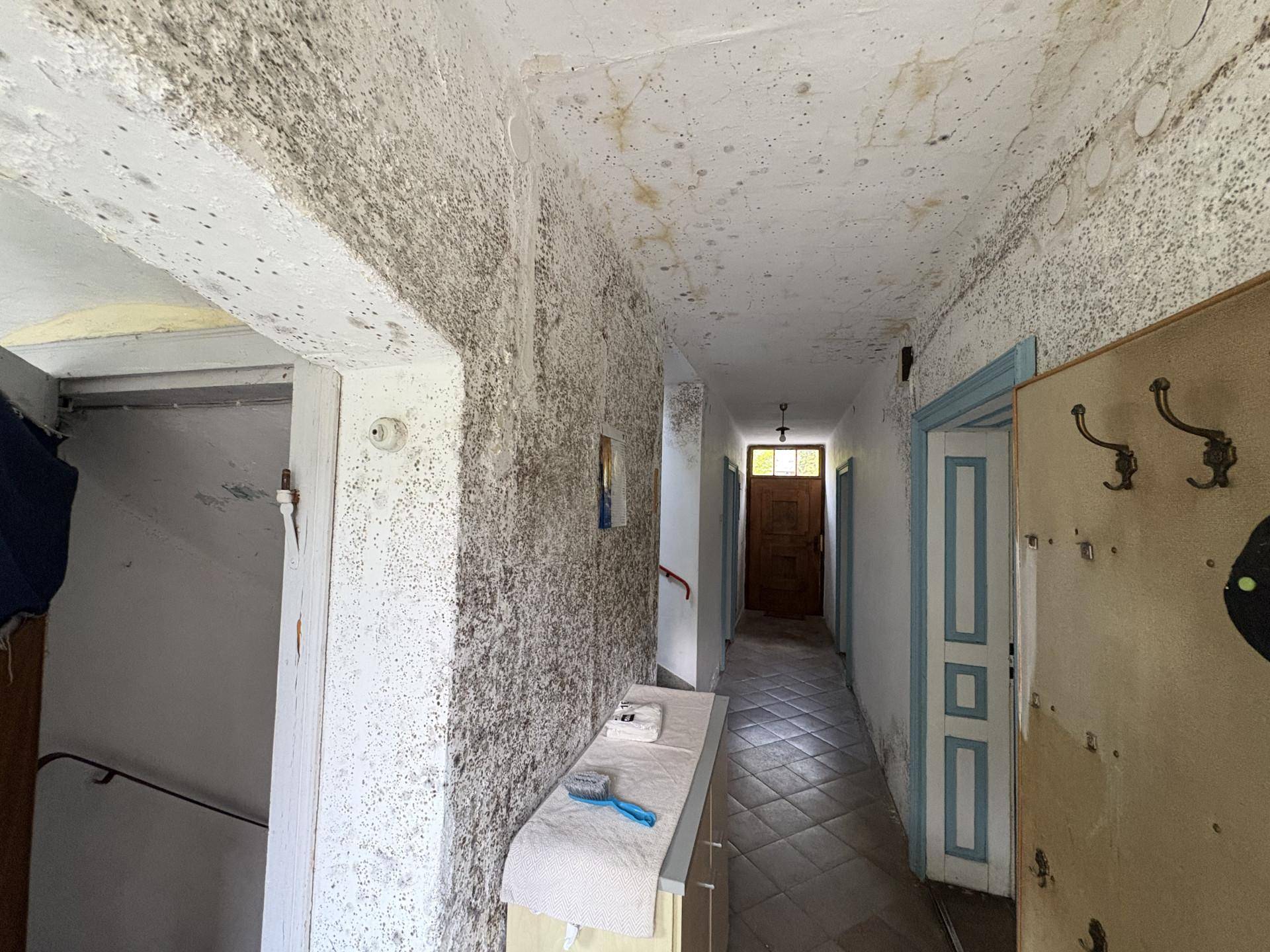 Langer Flur mit Fliesenboden, mehreren Türen und sichtbaren Feuchtigkeitsschäden an Wänden und Decke.