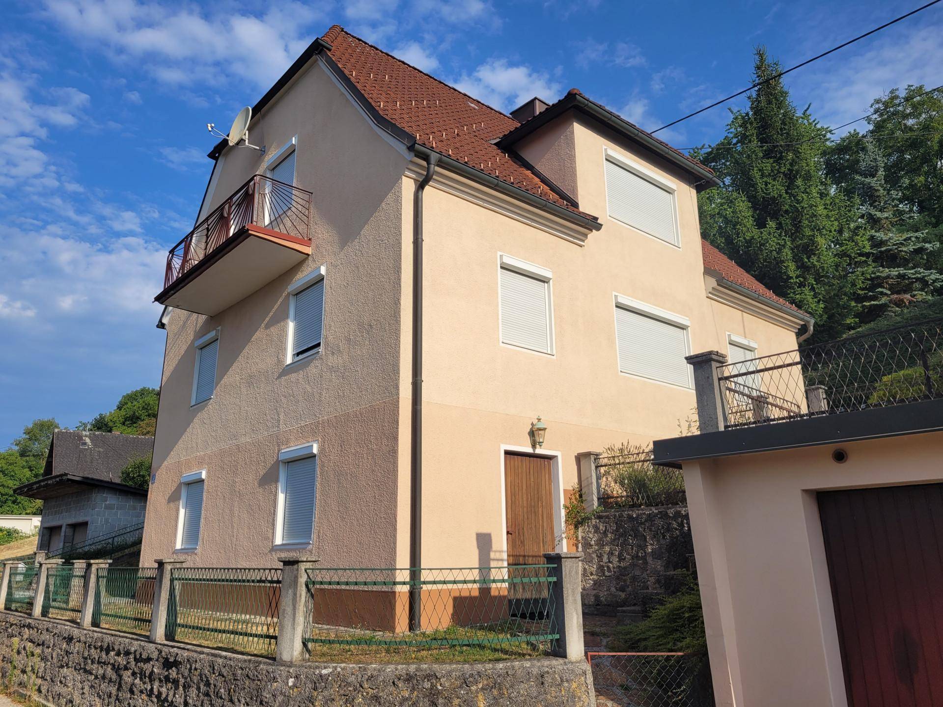 Vorderansicht des Hauses mit Eingangsbereich, Balkon und umliegender Bepflanzung bei Sonnenschein.
