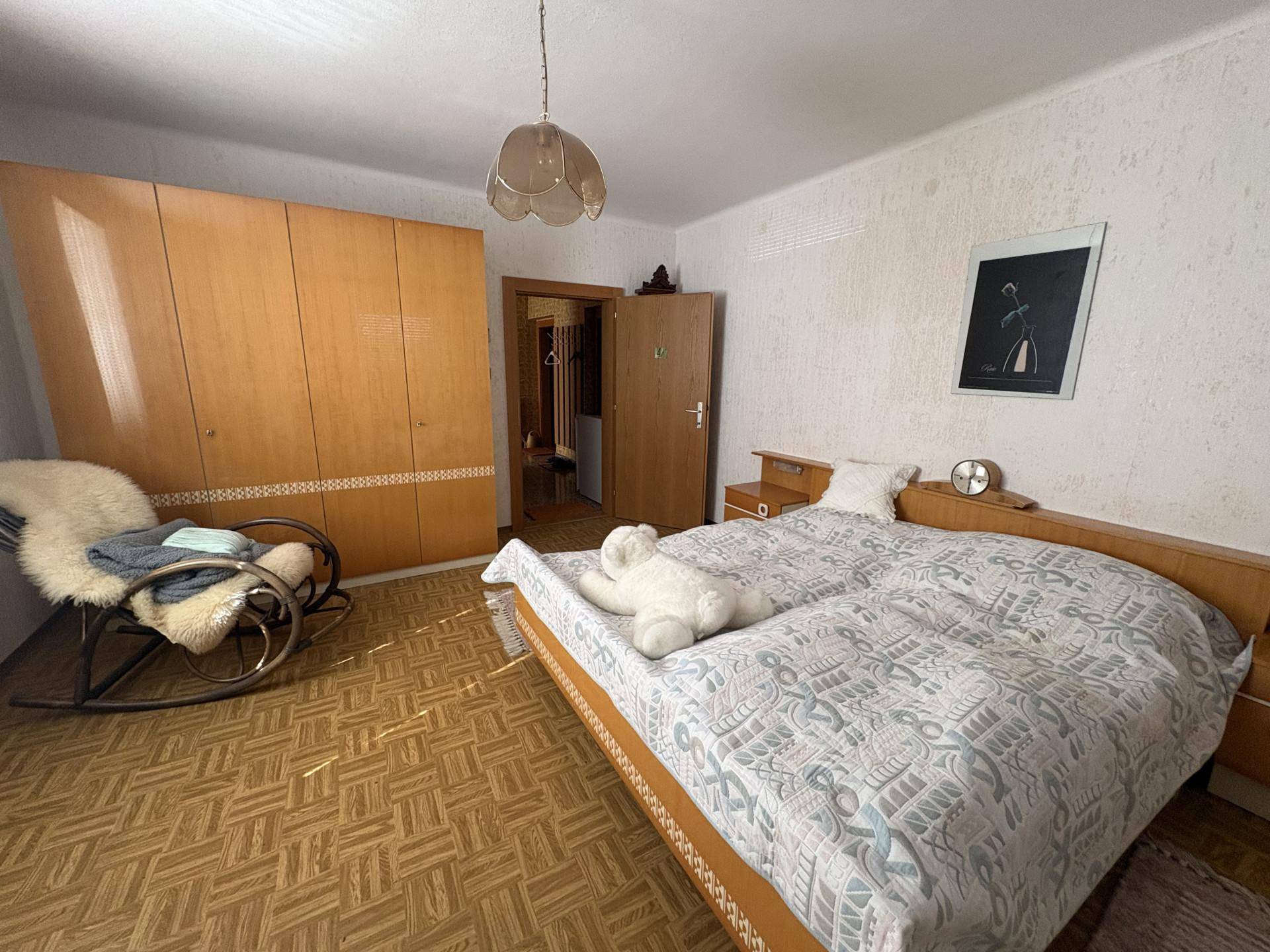Schlafzimmer mit Doppelbett, großem Holzschrank und einem Schaukelstuhl in warmer Atmosphäre.