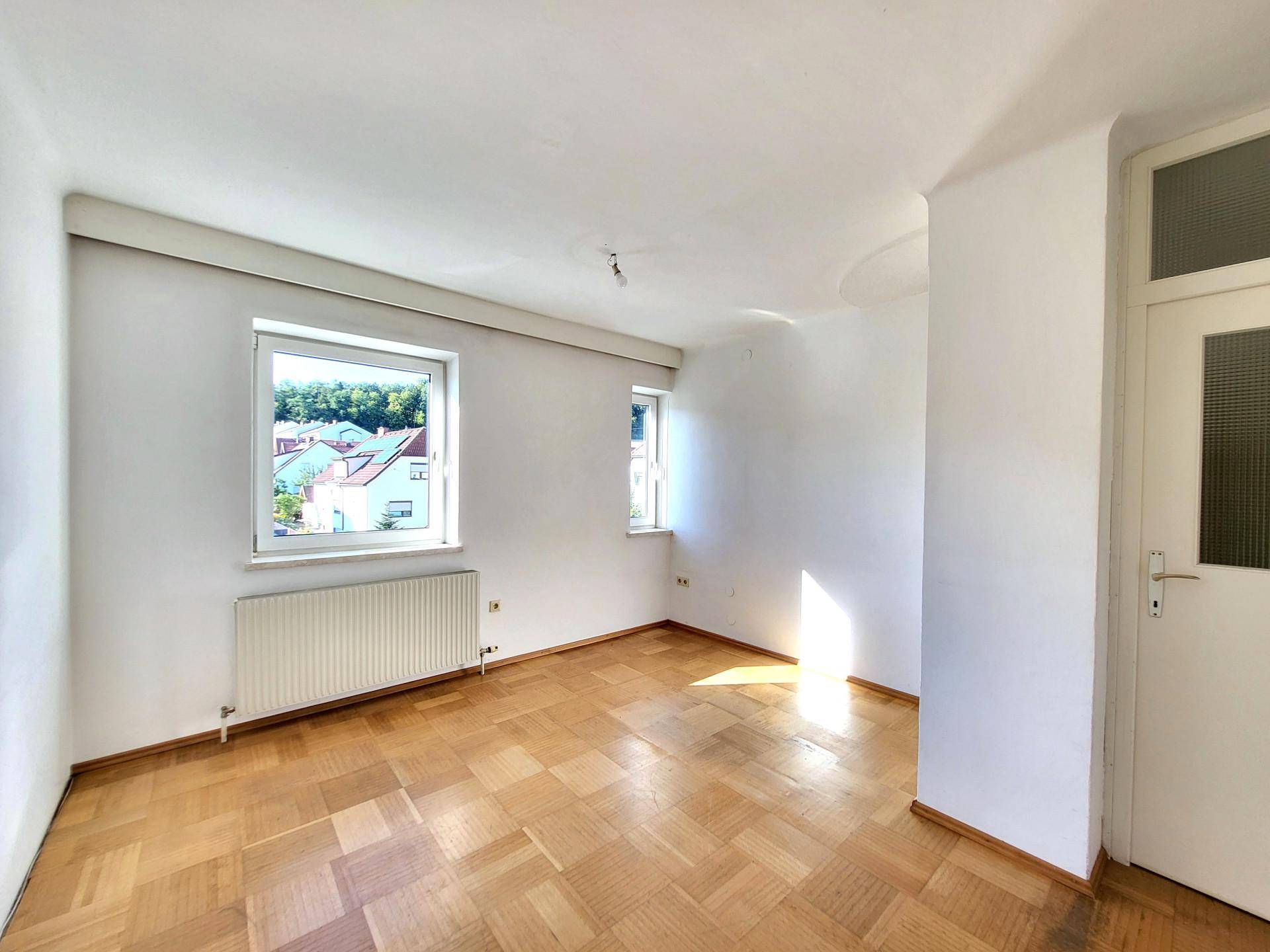 Heller Wohnraum mit Fischgrätparkettboden, weißen Wänden und zwei Fenstern für viel Tageslicht.