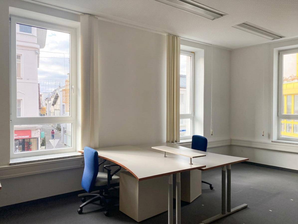 Helles Büro mit zwei Arbeitsplätzen, großen Fenstern und Blick auf die Straße, ausgestattet mit dunklem Teppichboden.
