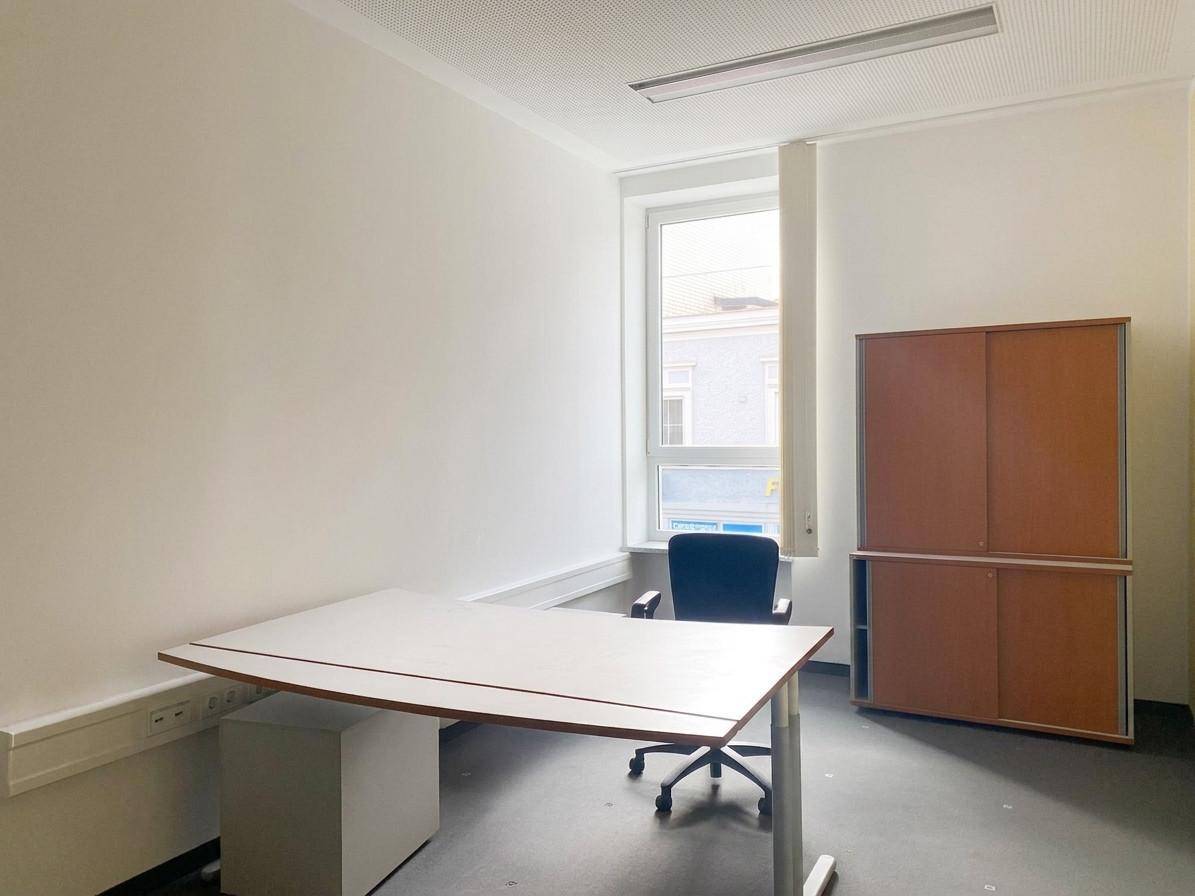 Einzelner Arbeitsplatz in einem hellen Büro mit großem Fenster und einem praktischen Holzschrank, ideal für konzentriertes Arbeiten.