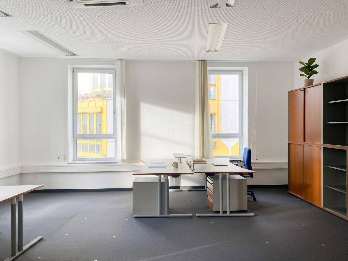 Geräumiges Büro mit mehreren Arbeitsplätzen, großen Fenstern und einem Holzschrank, gut beleuchtet durch Tageslicht.