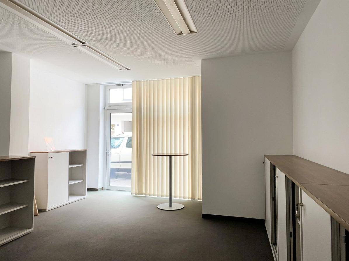 Heller Büroraum mit einer Glastür, vertikalen Jalousien und niedrigen Schränken, bietet direkten Zugang nach draußen oder zum Flur.