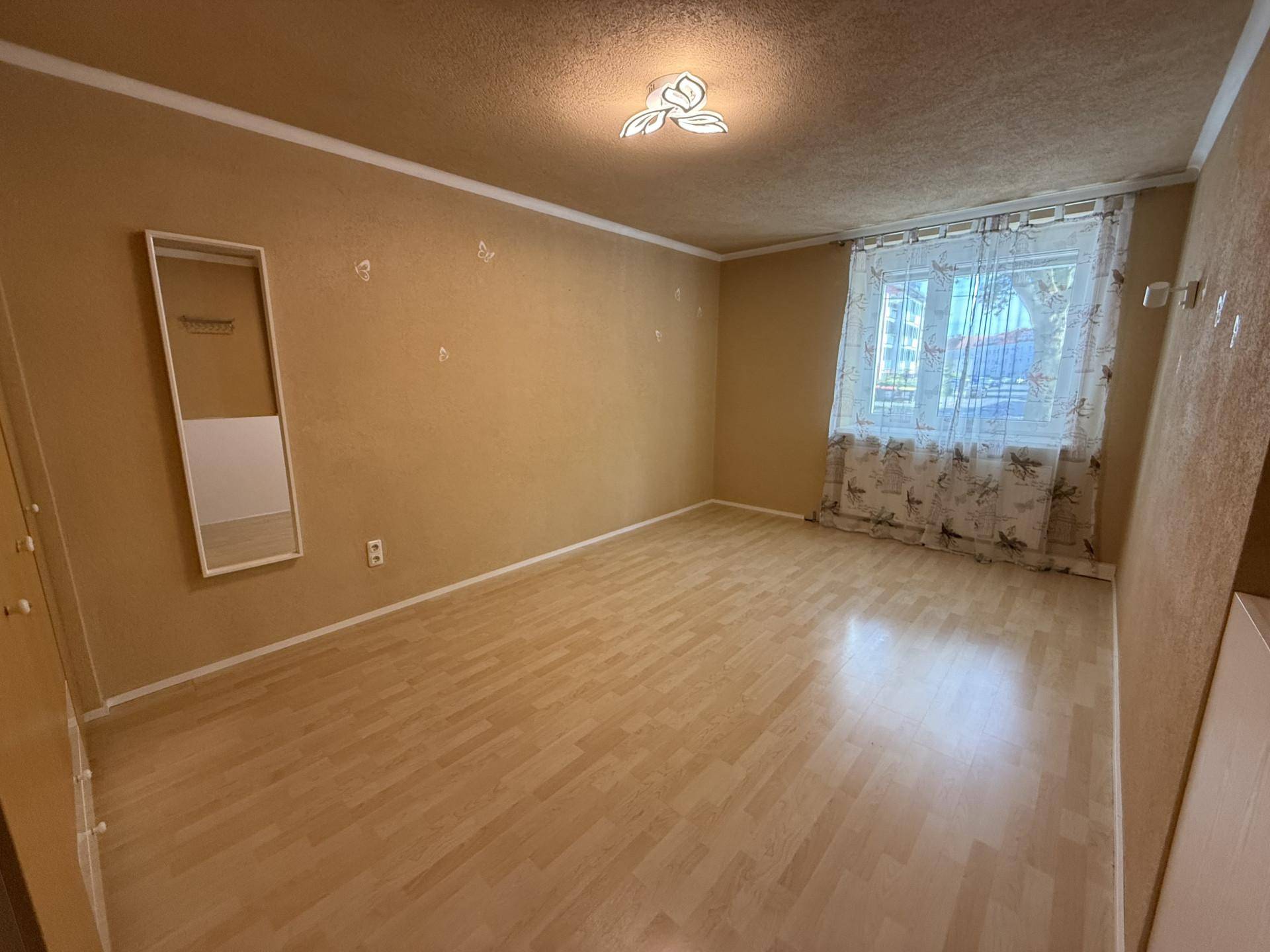 Leeres Schlafzimmer mit Laminatboden, beigen Wänden und einem Fenster mit gemusterten Vorhängen.