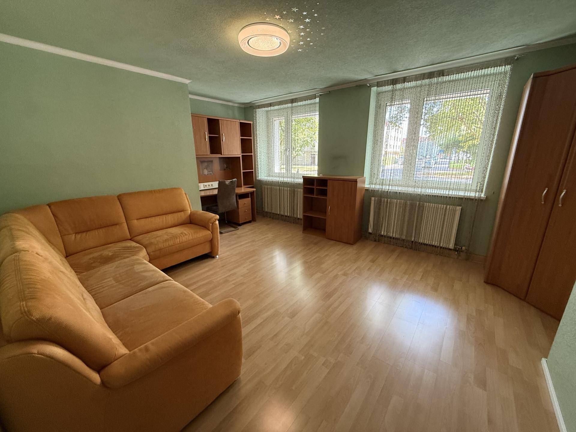 Helles Zimmer mit grünen Wänden, Laminatboden, einem Ecksofa und einem Arbeitsbereich am Fenster.